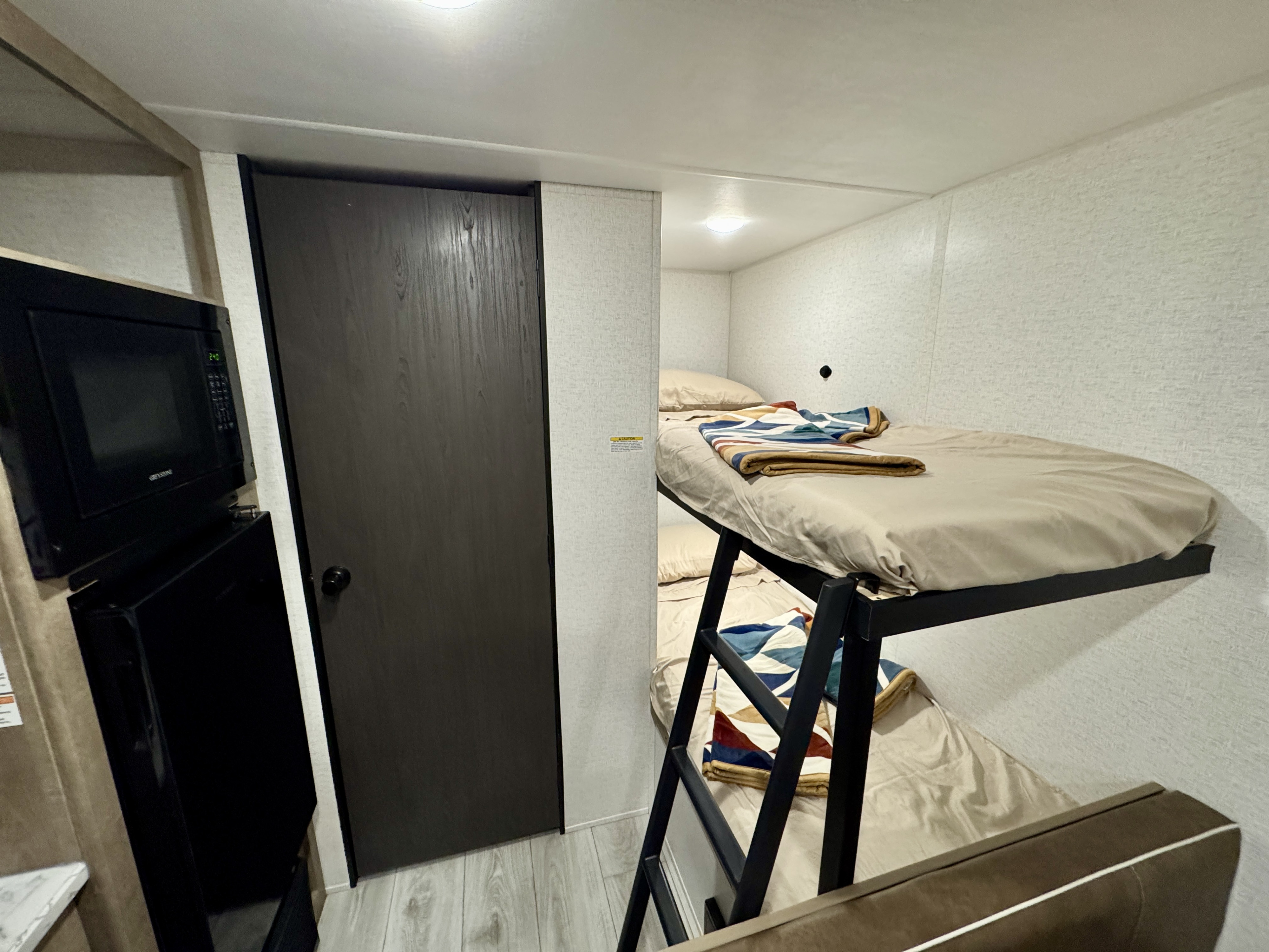 RV rental interior view of bunk beds with ladder, light bedding, a microwave, and a dark wood door in a compact space.. Keystone RV Springdale Mini 2025
