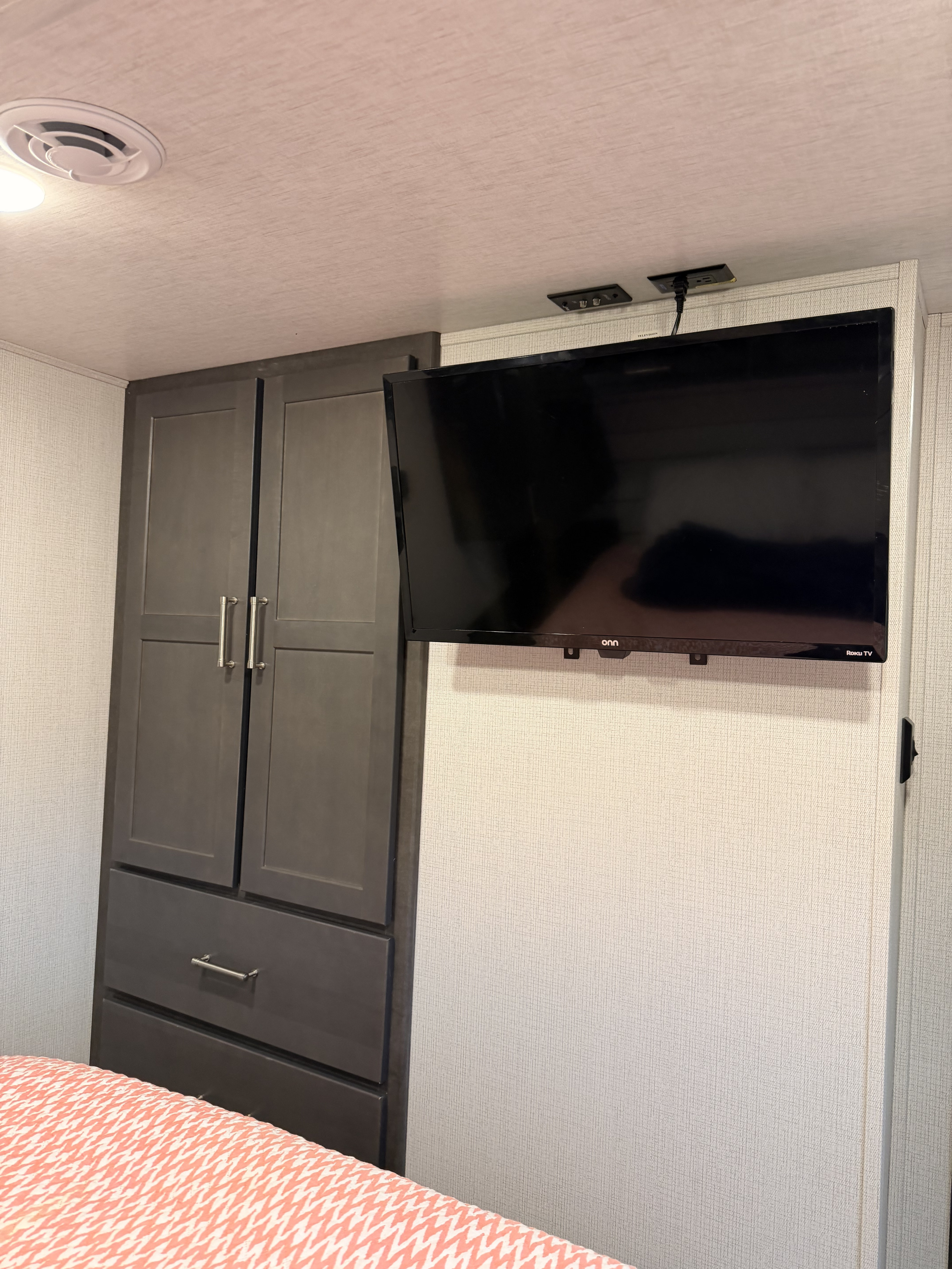 Cozy rv rental bedroom with dark cabinets, mounted Onn Roku TV, and patterned comforter. Features an air vent and power outlets.. Keystone RV Cougar Sport 2025