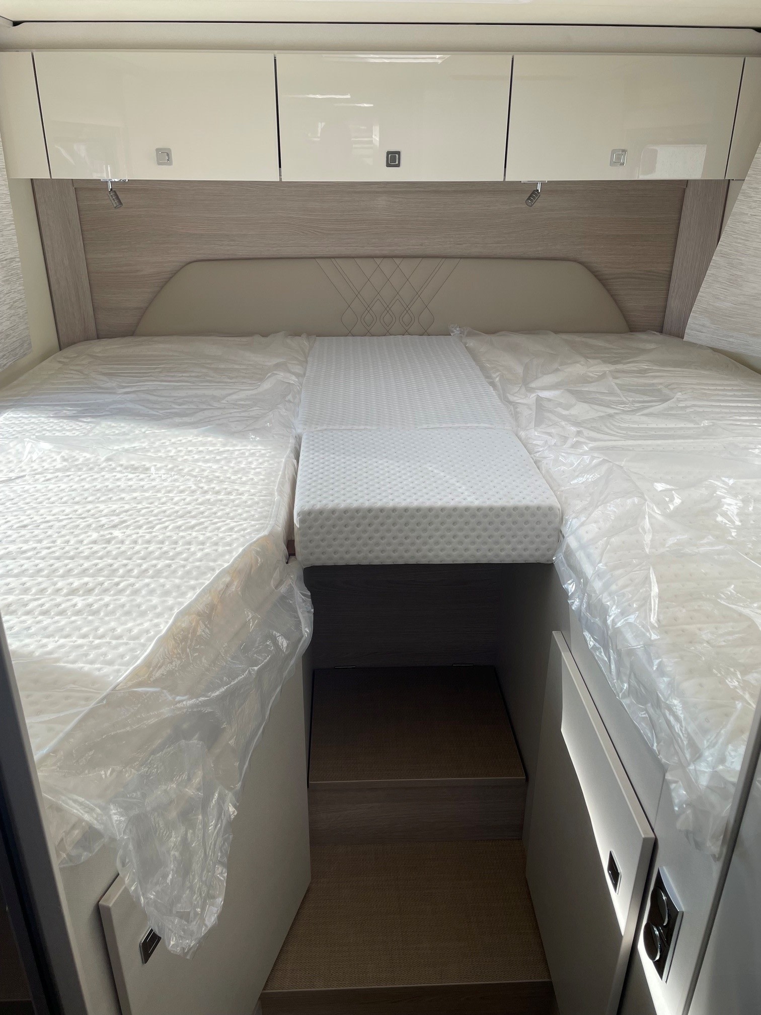 This RV rental bedroom features a clean, new mattress setup, overhead cabinets, and convenient steps below for storage access.. BURSTNER T690 2023