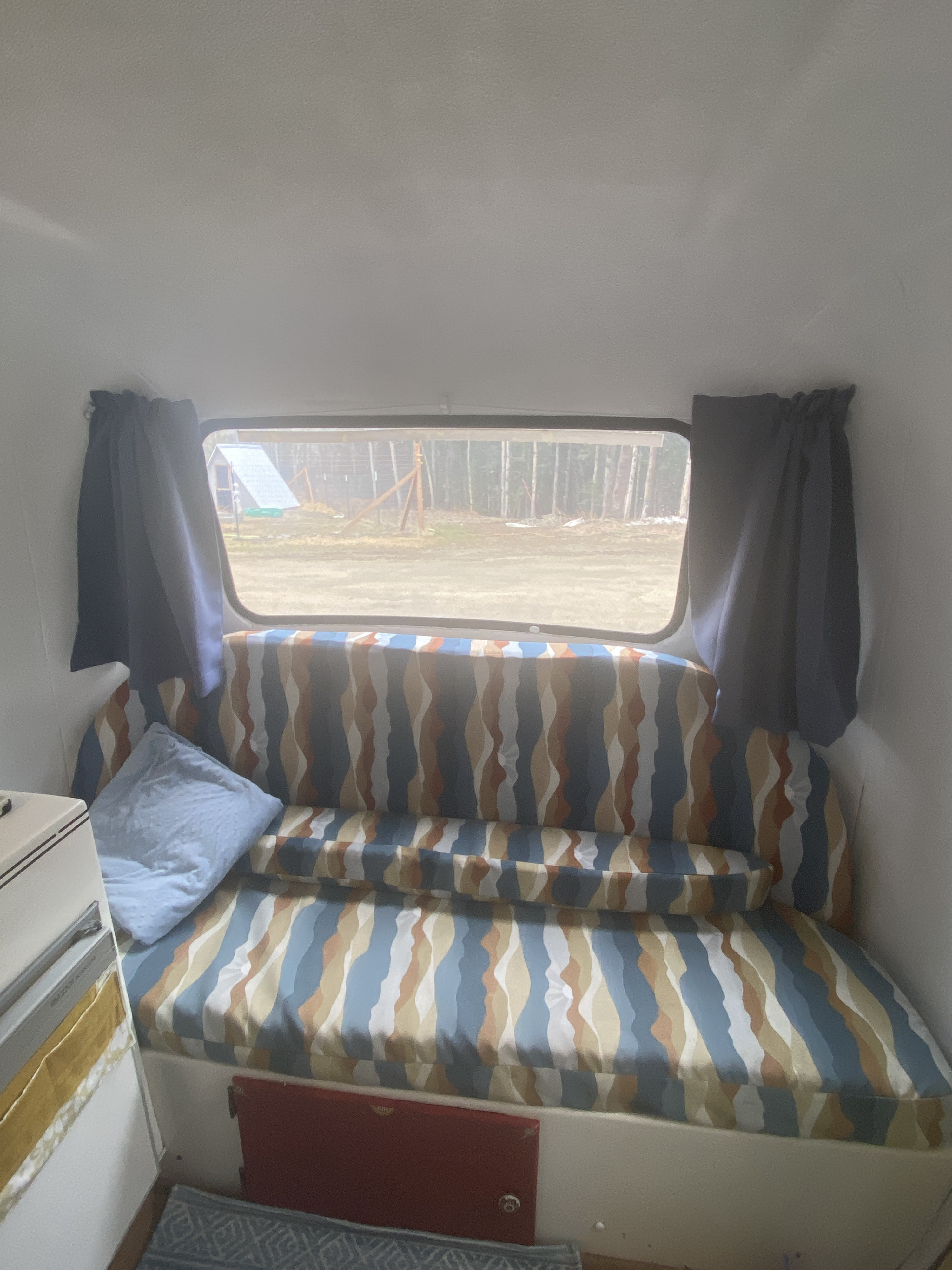 Cozy rv rental interior featuring a patterned bench/bed, window view of trees, and blue pillow. Ideal for comfortable travel.. Scamp 13' 1987