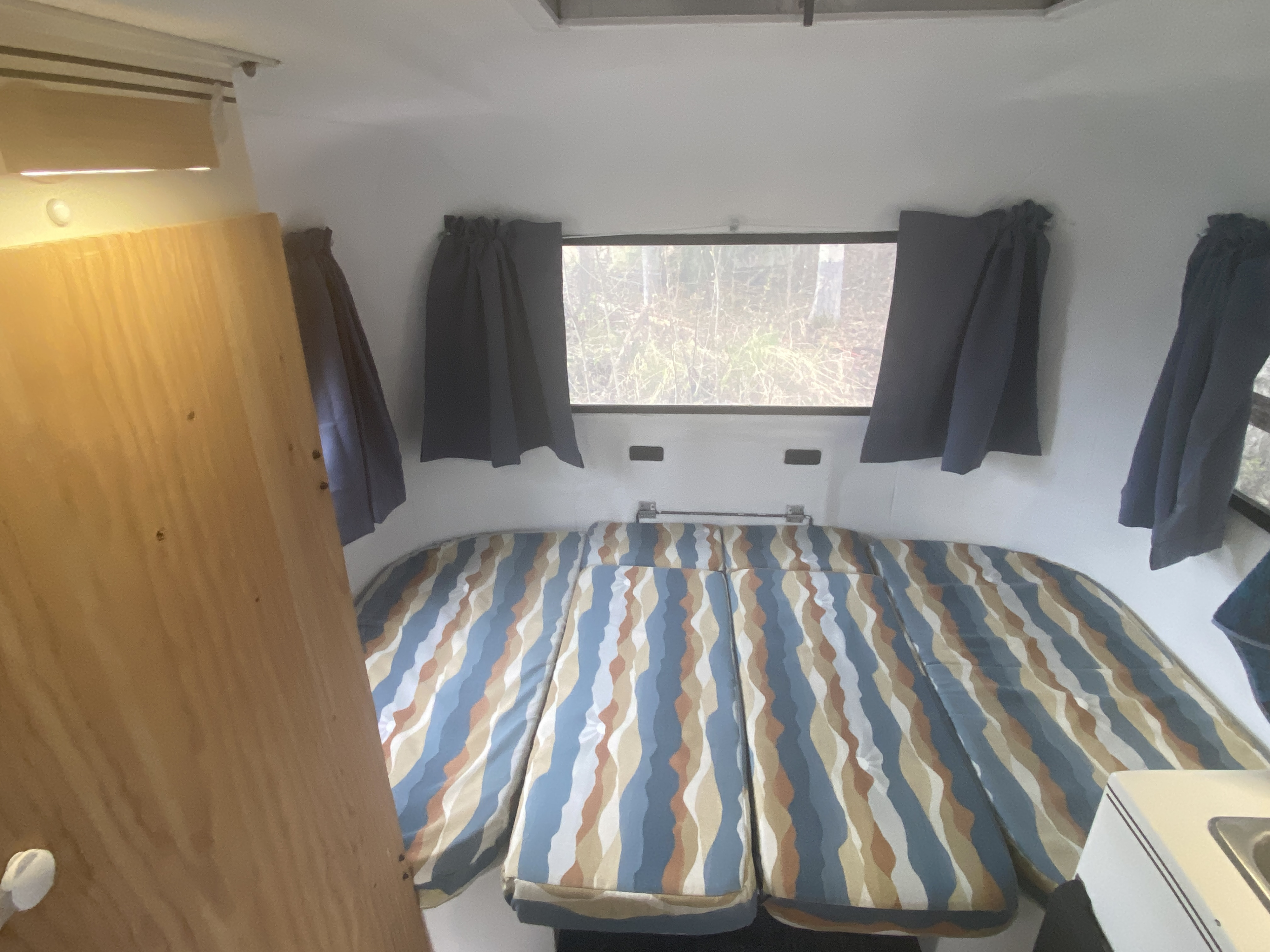 RV rental interior: A cozy sleeping area with colorful striped cushions, a large window showcasing nature, and blue curtains.. Scamp 13' 1987