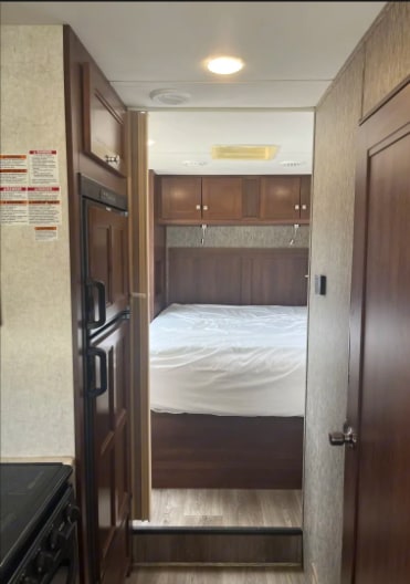 RV rental interior: A compact bedroom with a white bed and overhead cabinets, viewed from a narrow hallway with a kitchen on the left.. Forest River Sunseeker 2018