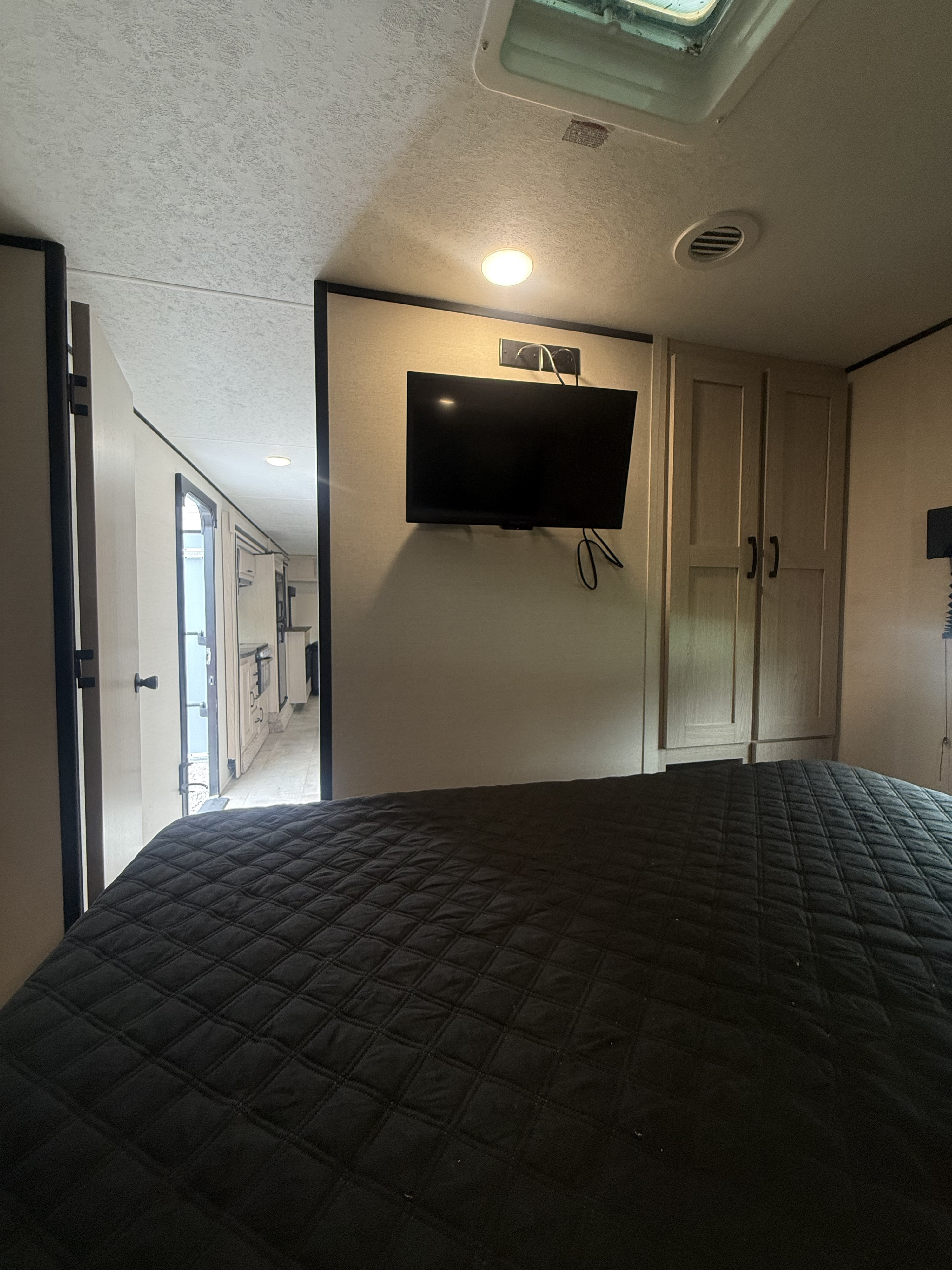 RV rental bedroom featuring a dark quilted bedspread, wall-mounted TV, wardrobe, and open door leading to the interior.. Coachmen Apex 2021