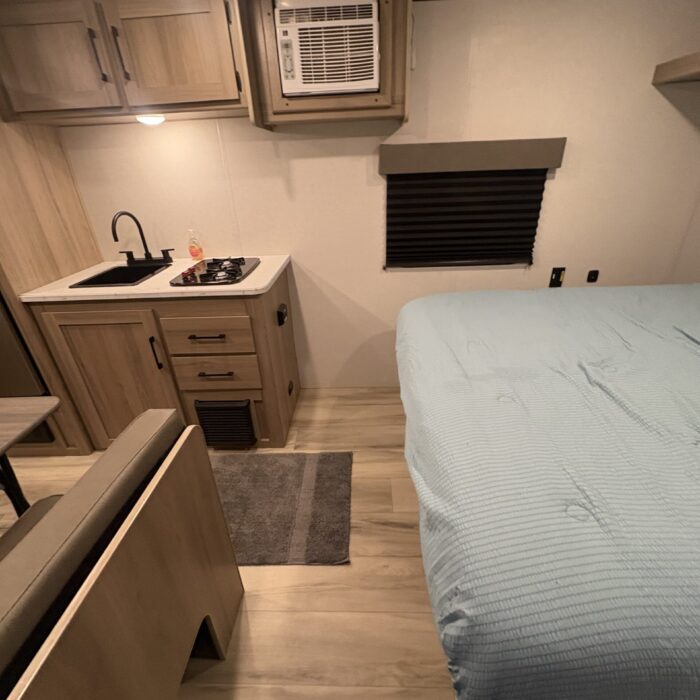 A bright interior view of an RV rental showing a compact kitchen with a sink and stove, an air conditioning unit, and a bed with a light blue cover.. Keystone RV Coleman 2025