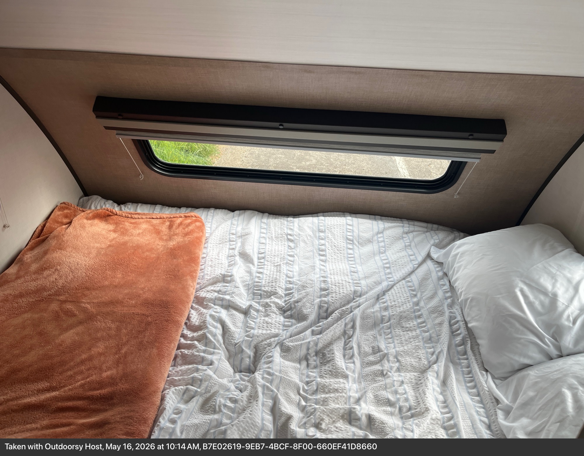 Cozy RV rental bedroom features a white striped bed, orange blanket, and window with a view of green grass and pavement.. Forest River R pod 2021