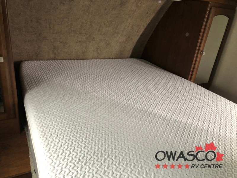 Inside an RV rental: a cozy bed with a textured white mattress cover, surrounded by wood trim and wall paneling.. Coachmen Freedom Express 312BHDS Travel Trailer 2017