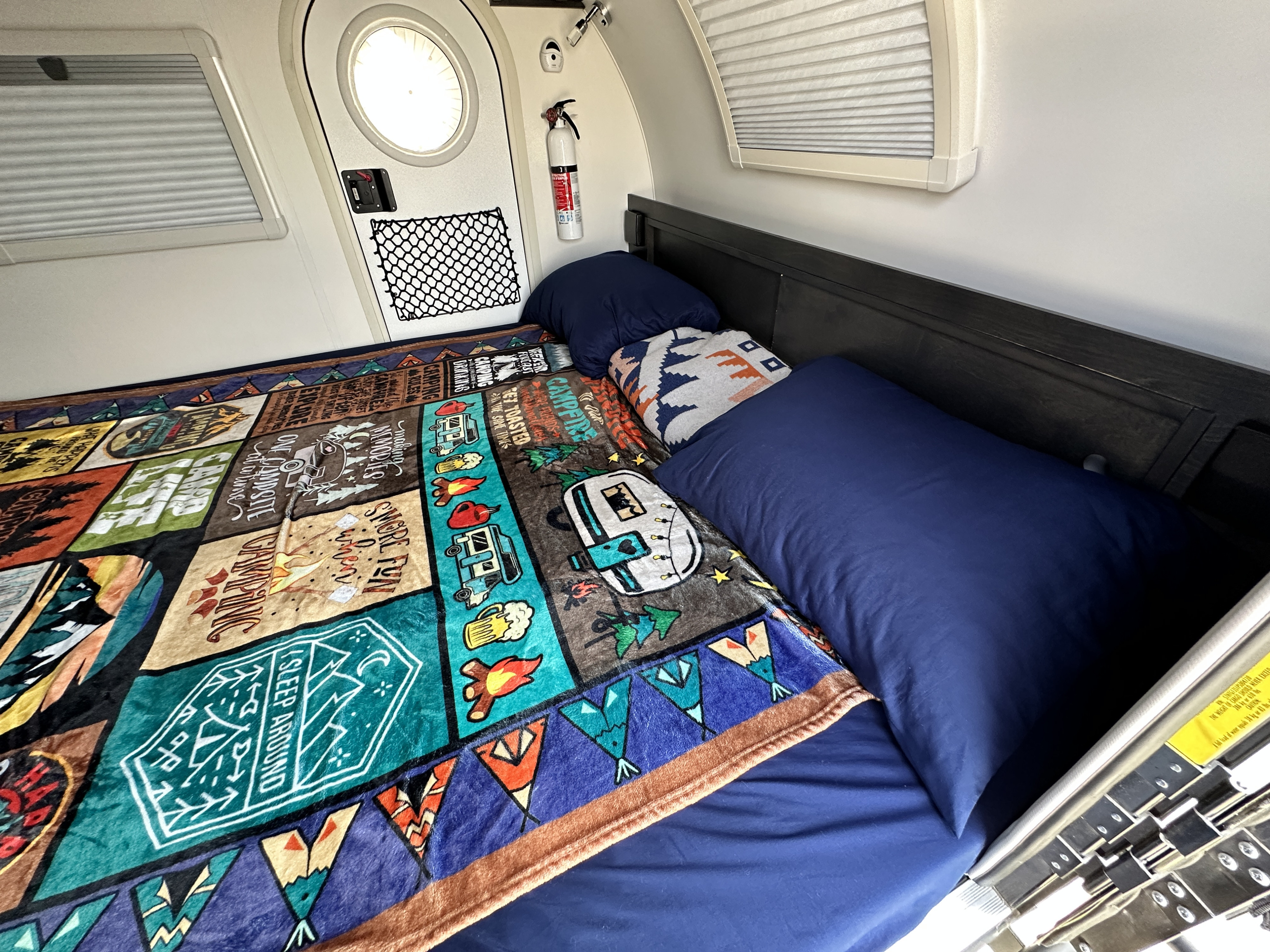 Interior of an RV rental showing a bed with blue linens & a colorful camping blanket, fire extinguisher, and windows.. nuCamp T@G 2023