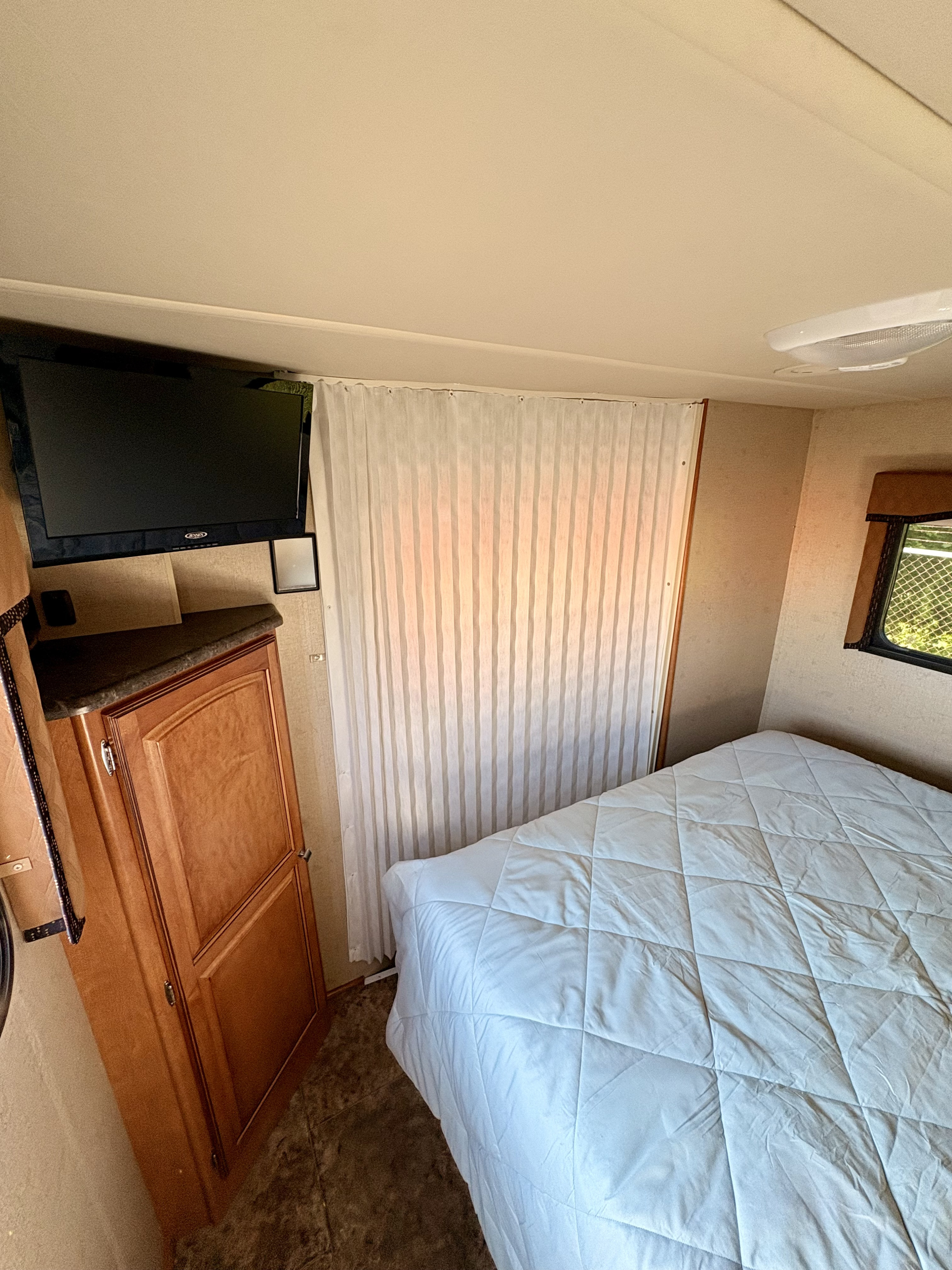 This RV rental bedroom features a white quilted bed, wall-mounted TV, wood cabinet, and a window with a sheer curtain.. Winnebago Access 2013