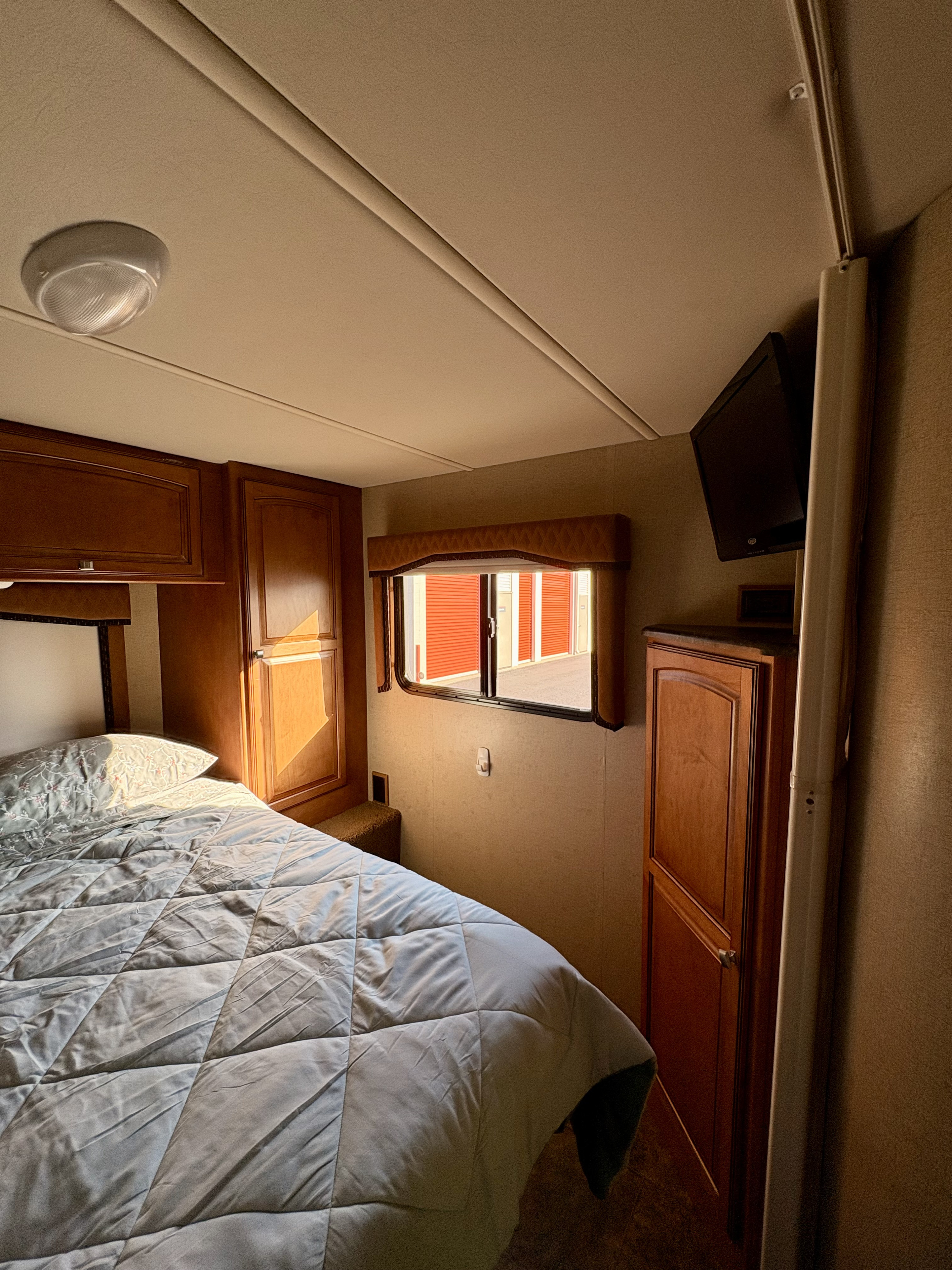 RV rental bedroom with comfy bed, wood cabinets, wall-mounted TV, and a window showing storage units.. Winnebago Access 2013