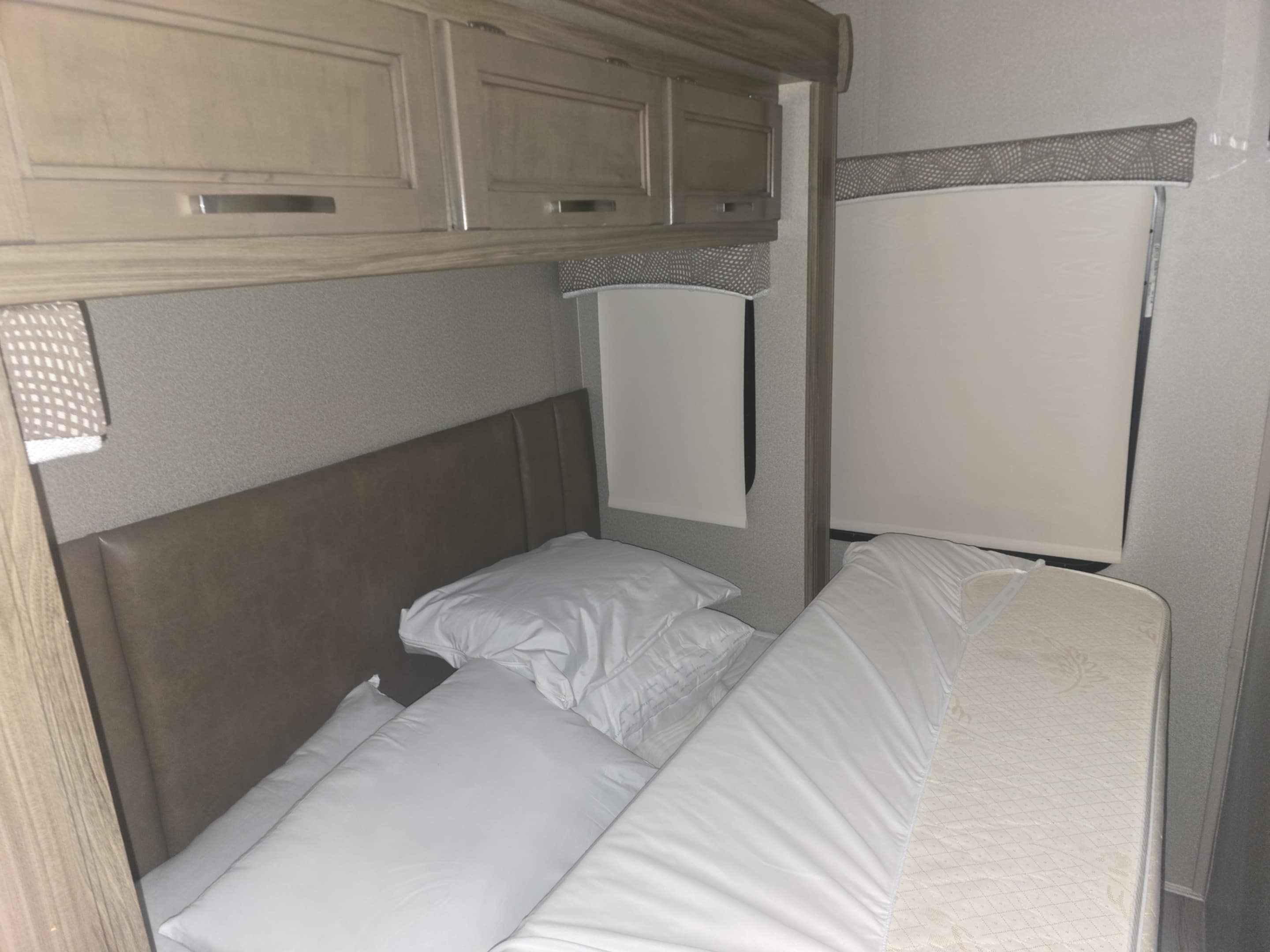 RV rental bedroom with a bed, white linens, padded headboard, wood overhead cabinets, and light-colored window blinds.. Entegra Coach Vision 2019