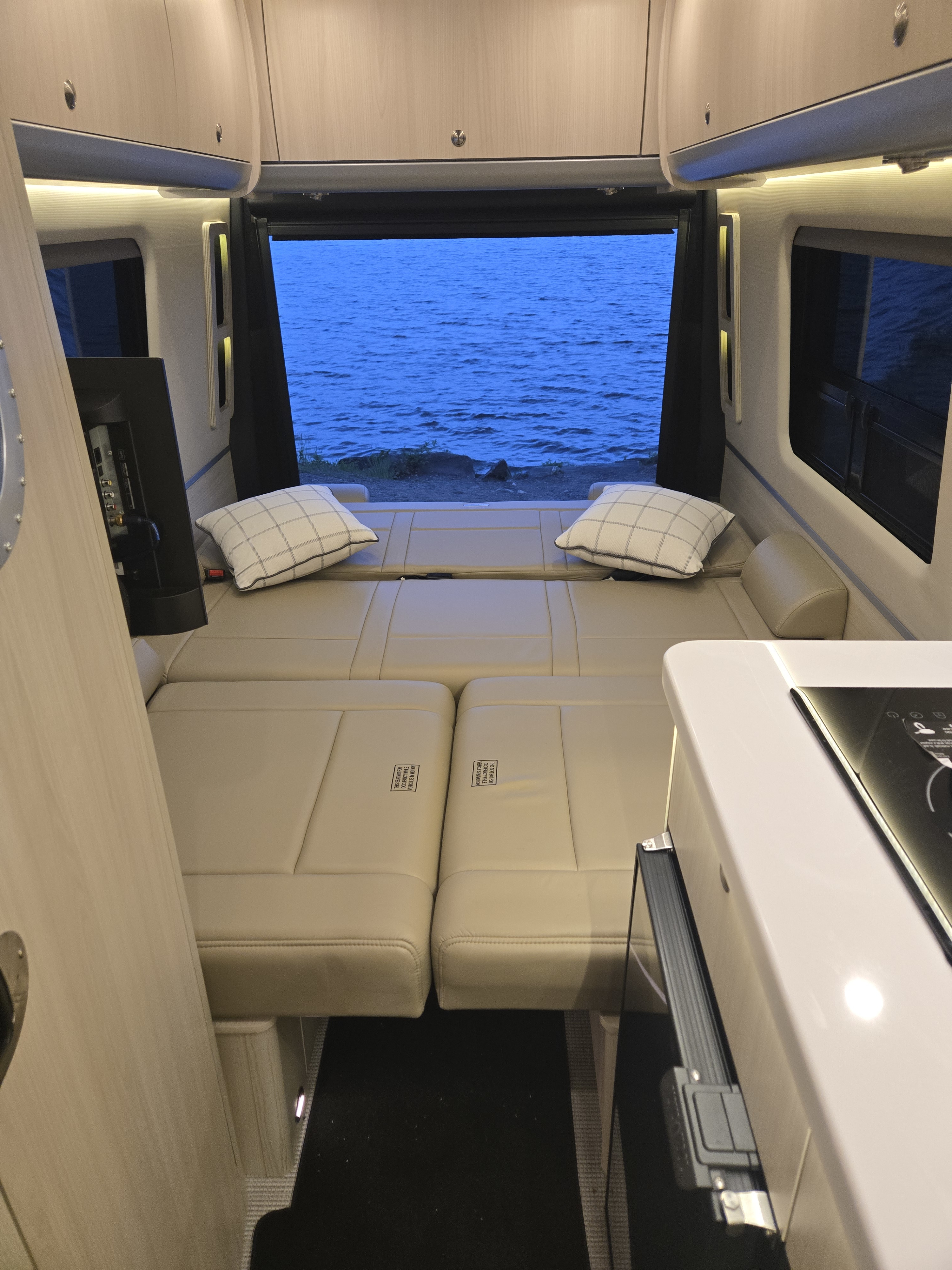 Interior of a modern rv rental shows a cream upholstered bed, compact kitchen counter, and open doors revealing a tranquil blue lake.. Airstream Interstate 2026