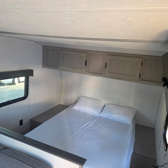 RV rental bedroom with a neat white bed, overhead cabinets, and a window, offering comfortable sleeping space in a compact setup.. Clipper 5K 2024