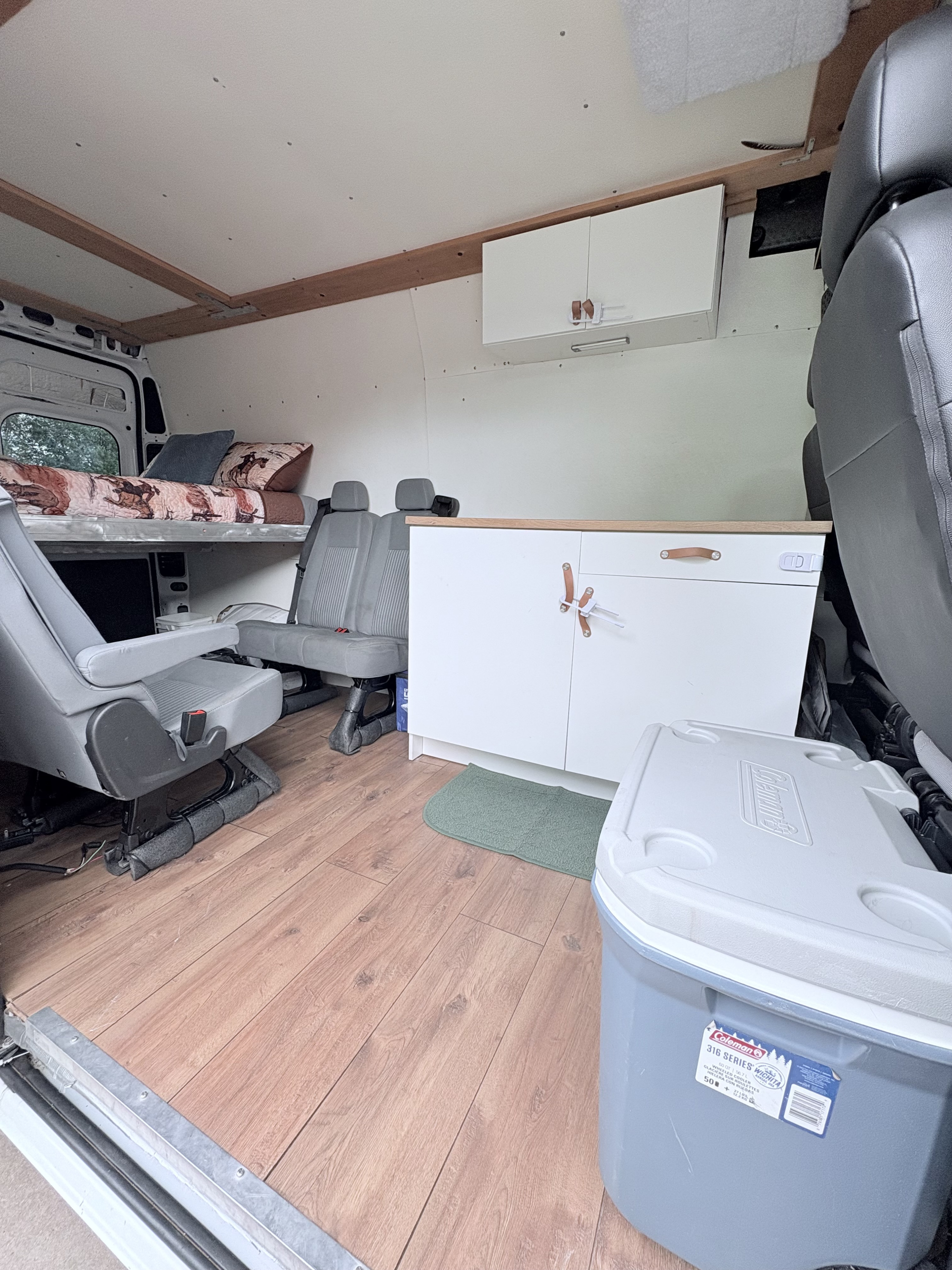Inside an rv rental: custom van interior with an elevated bed, gray seating, and storage, including a Coleman cooler.. Dodge 2500 Cargo Van High Roof 159" WB 2019