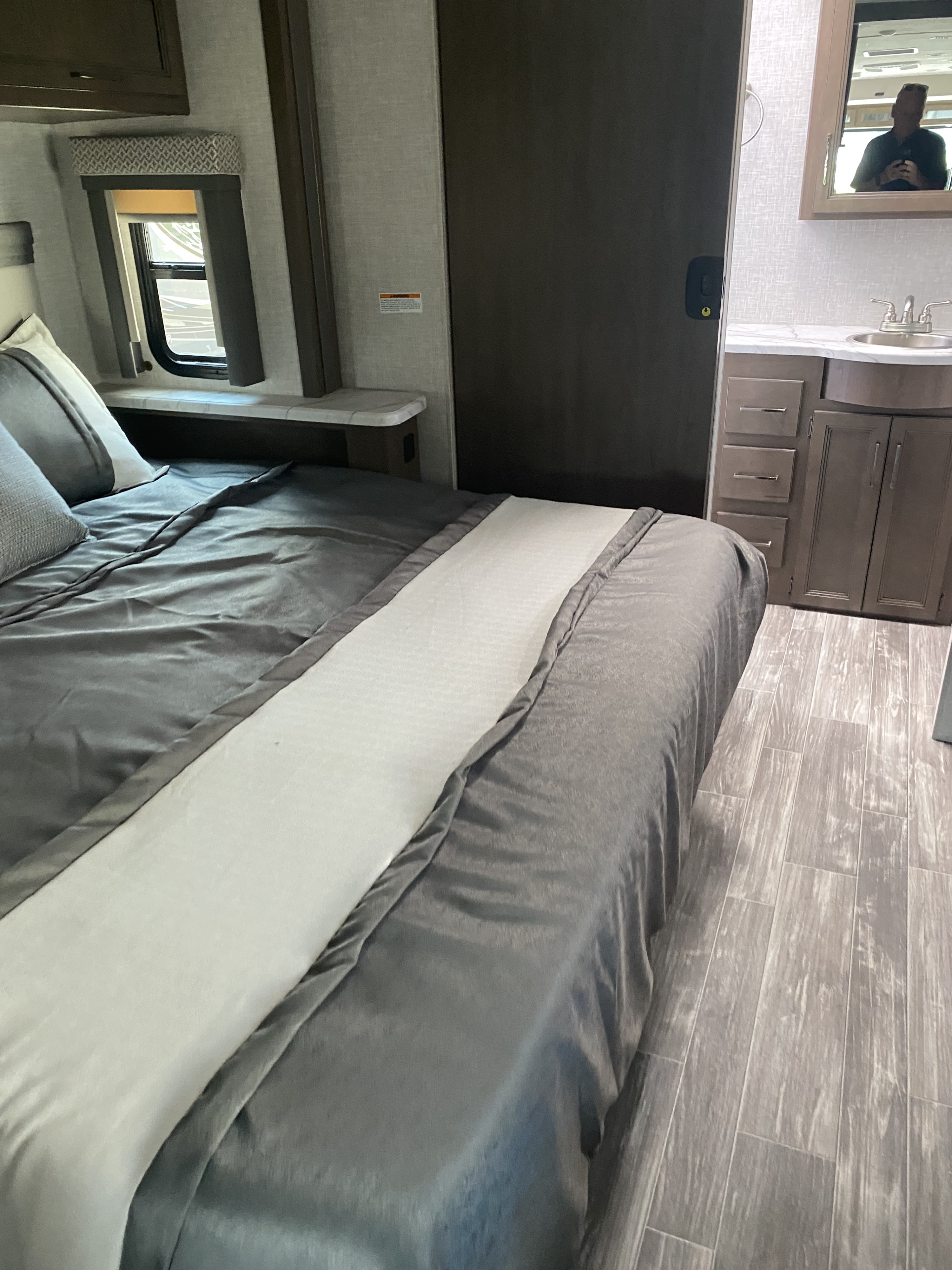 Cozy RV rental bedroom featuring a grey bed with white runner, window, and a connected vanity and sink. Modern grey interior design.. Thor Hurricane 2022