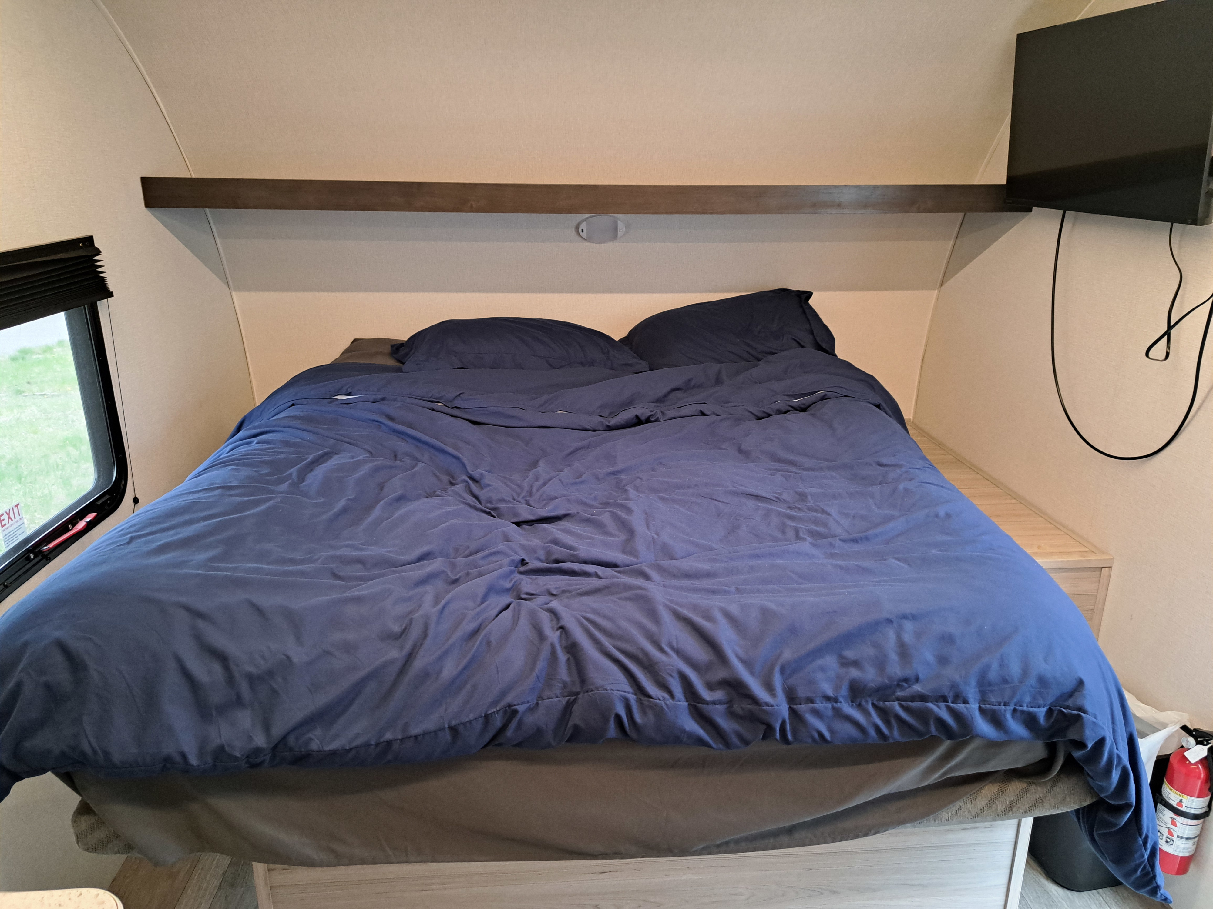 Comfortable RV rental bedroom with blue bed, overhead shelf, TV, and fire extinguisher for safety.. Heartland RVs Mallard 2025