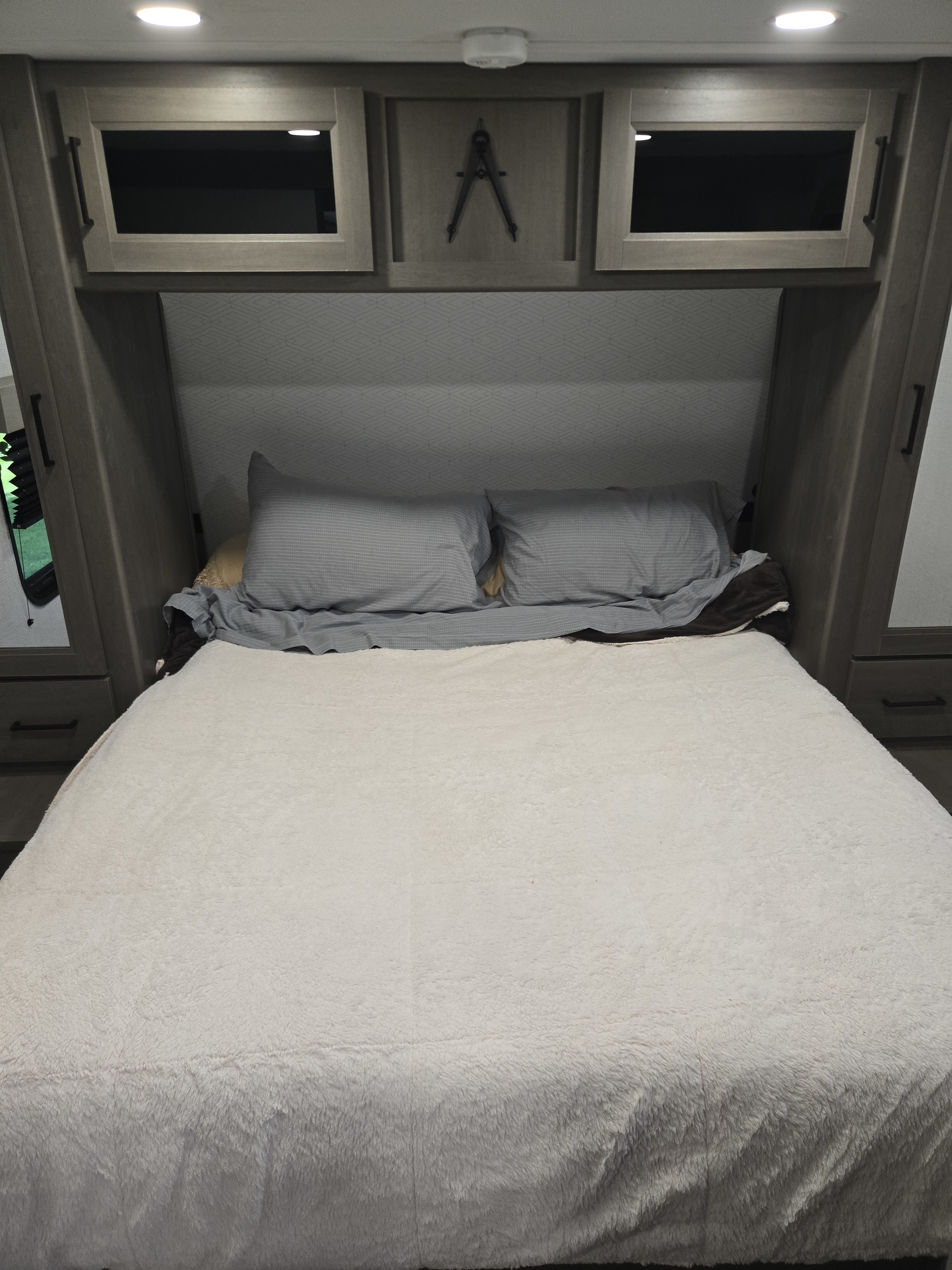 Inside a modern RV rental: a well-appointed bedroom with a soft bed, geometric headboard, and ample overhead storage.. Grand Design Transcend 2026
