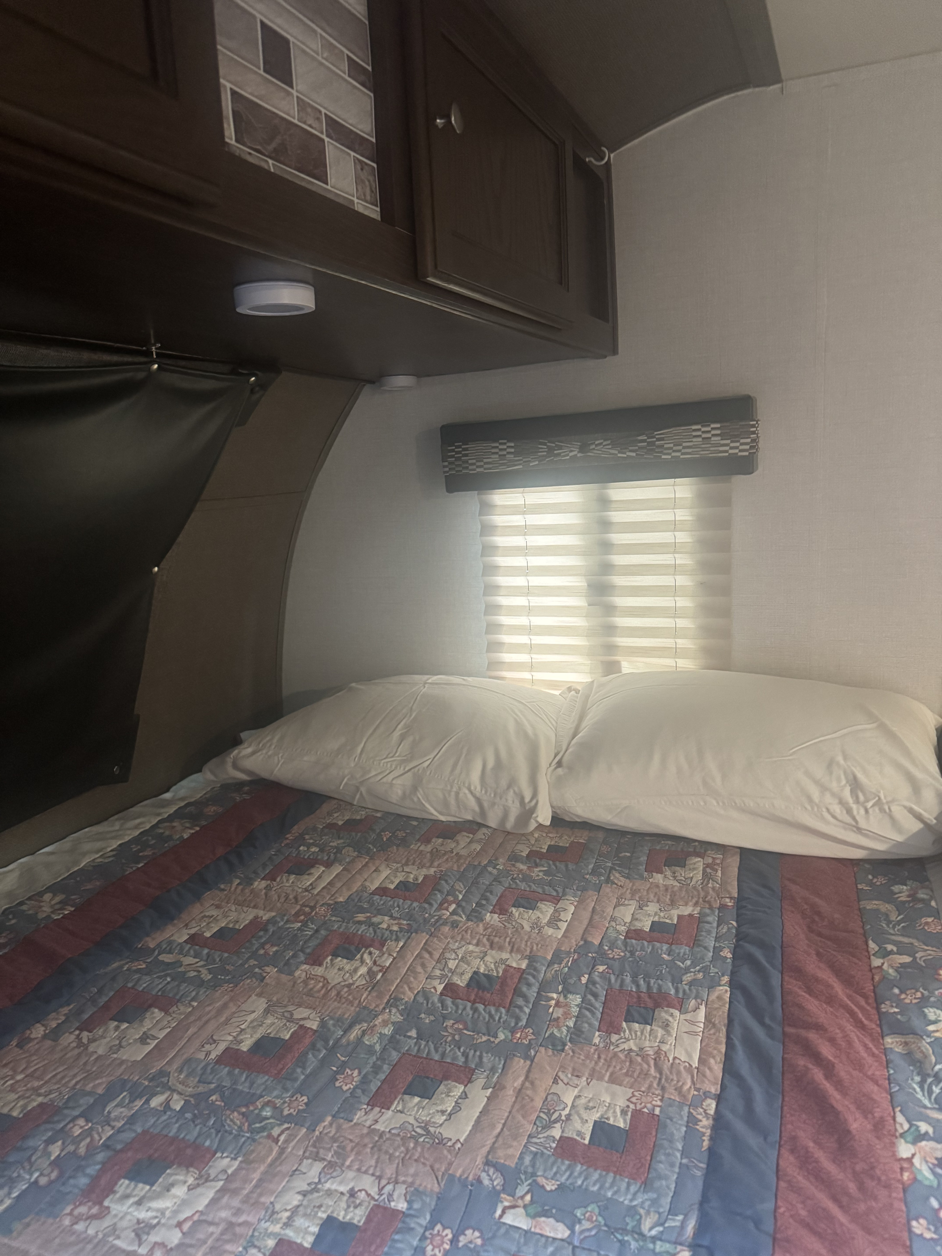 Inside an RV rental: a compact bedroom with a patterned quilt, pillows, window blinds, and overhead cabinets.. Forest River Palomino Palomini 2018