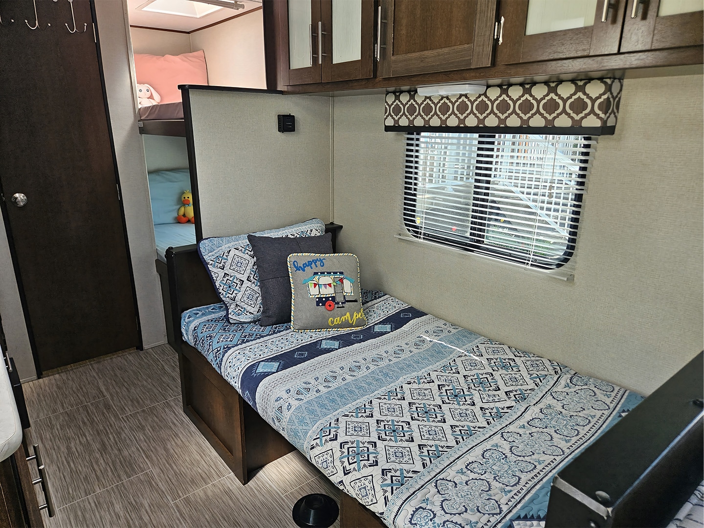 Cozy RV rental interior with a twin bed, patterned blue bedding, overhead cabinets, and bunk beds visible in the background.. Navi Navi Trailer 2019