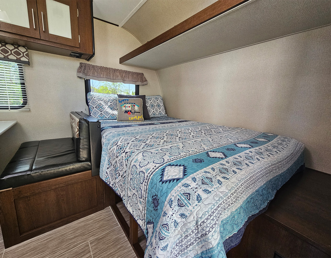 Cozy RV rental bedroom area with a patterned bedspread, 'Happy Camper' pillow, and a comfortable bench seat.. Navi Navi Trailer 2019
