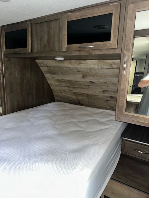 Inside an RV rental, this cozy bedroom features a comfortable bed, wood-look headboard, overhead cabinets, and mirrored nightstand.. Dutchmen Coleman Light 2019