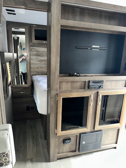 RV rental interior: Compact bedroom/living area with media console, bed, and mirrored closet reflecting a person.. Dutchmen Coleman Light 2019