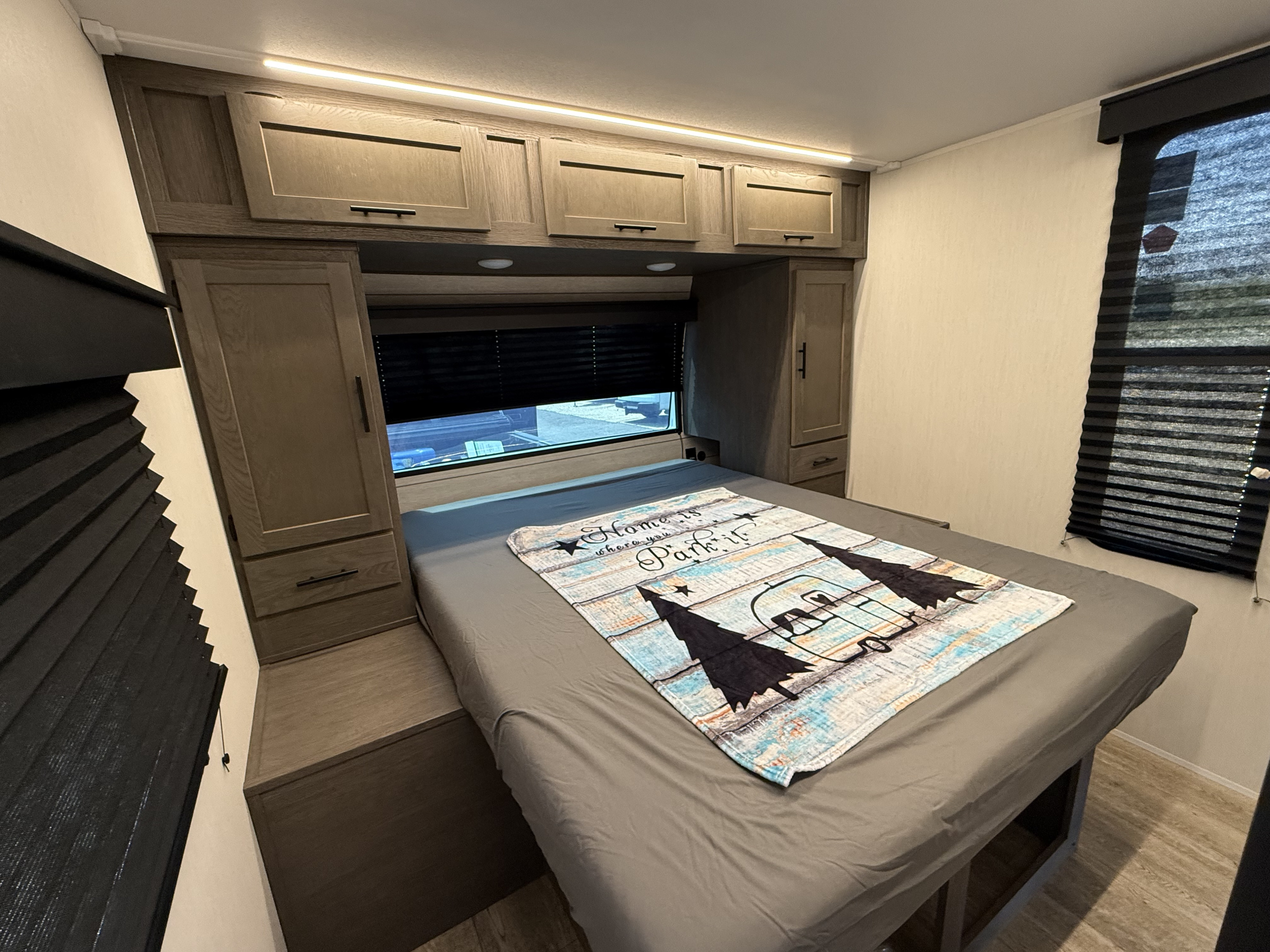 Cozy RV rental bedroom featuring a bed with a "Home is where you Park it" blanket, overhead cabinets, and window blinds.. Forest River Cherokee Alpha Wolf 2025