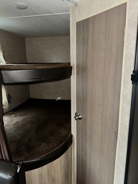 Inside an RV rental: two cozy bunk beds with dark bedding, next to a light wood door. Ideal sleeping for campers.. Coachmen SBX 261BH 2020