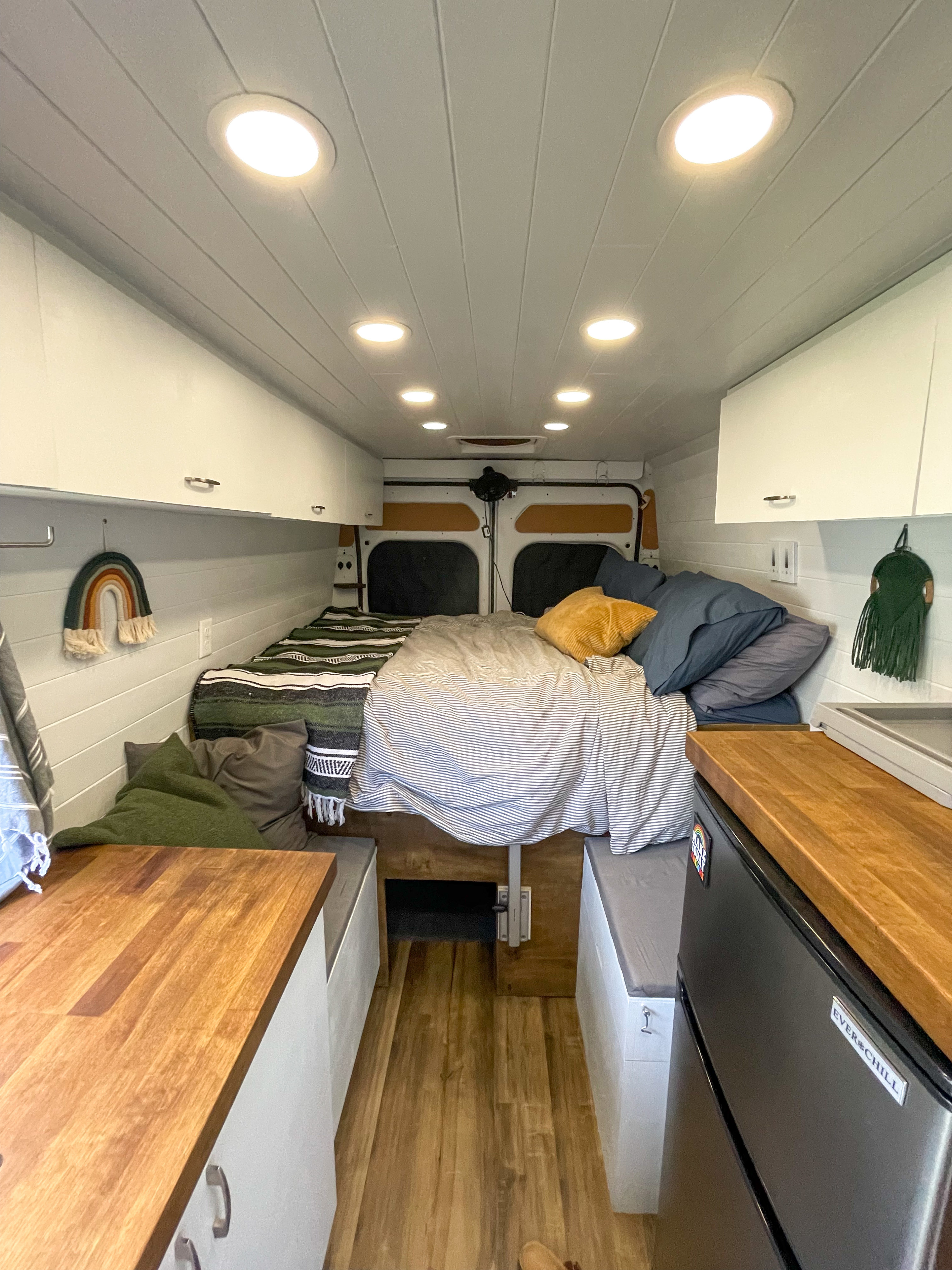 An RV rental interior, brightly lit, shows a bed with pillows, wooden counters, a fridge, and white overhead storage.. Dodge Promaster 2500 2019