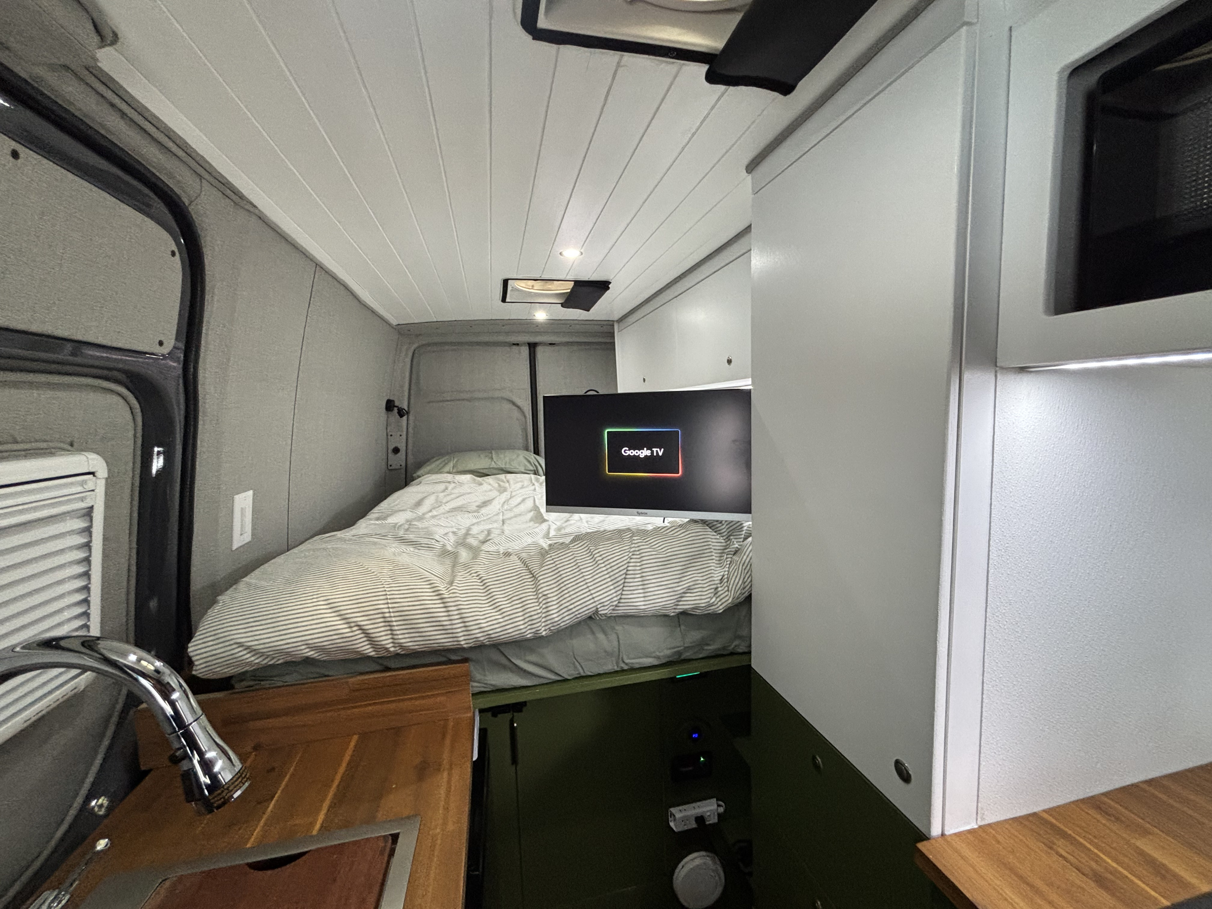 RV rental interior: Cozy bed, Google TV, and kitchen sink in a compact camper van space. Modern and functional design.. Mercedes-Benz Sprinter 2023