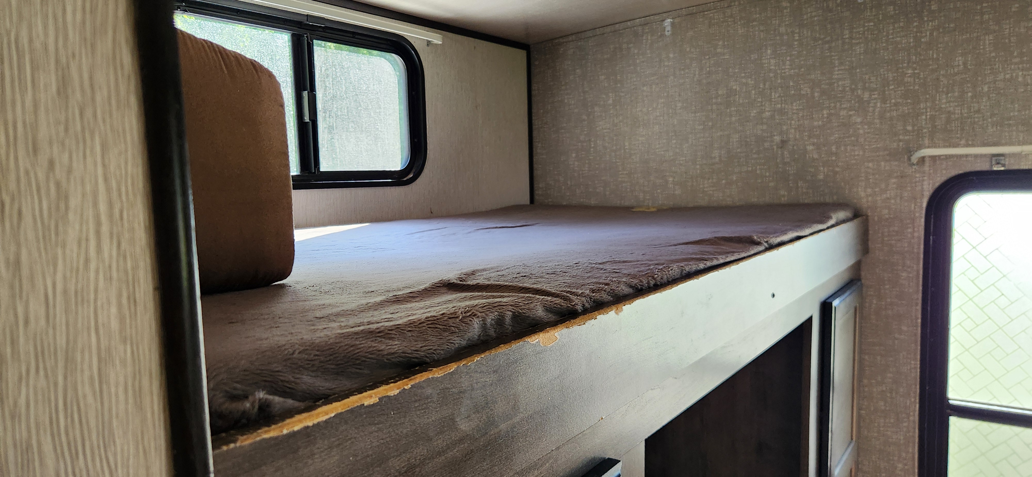 Inside an RV rental: a top bunk bed with a brown mattress cover and windows, showing the compact living space.. Heartland RVs Mallard 2019