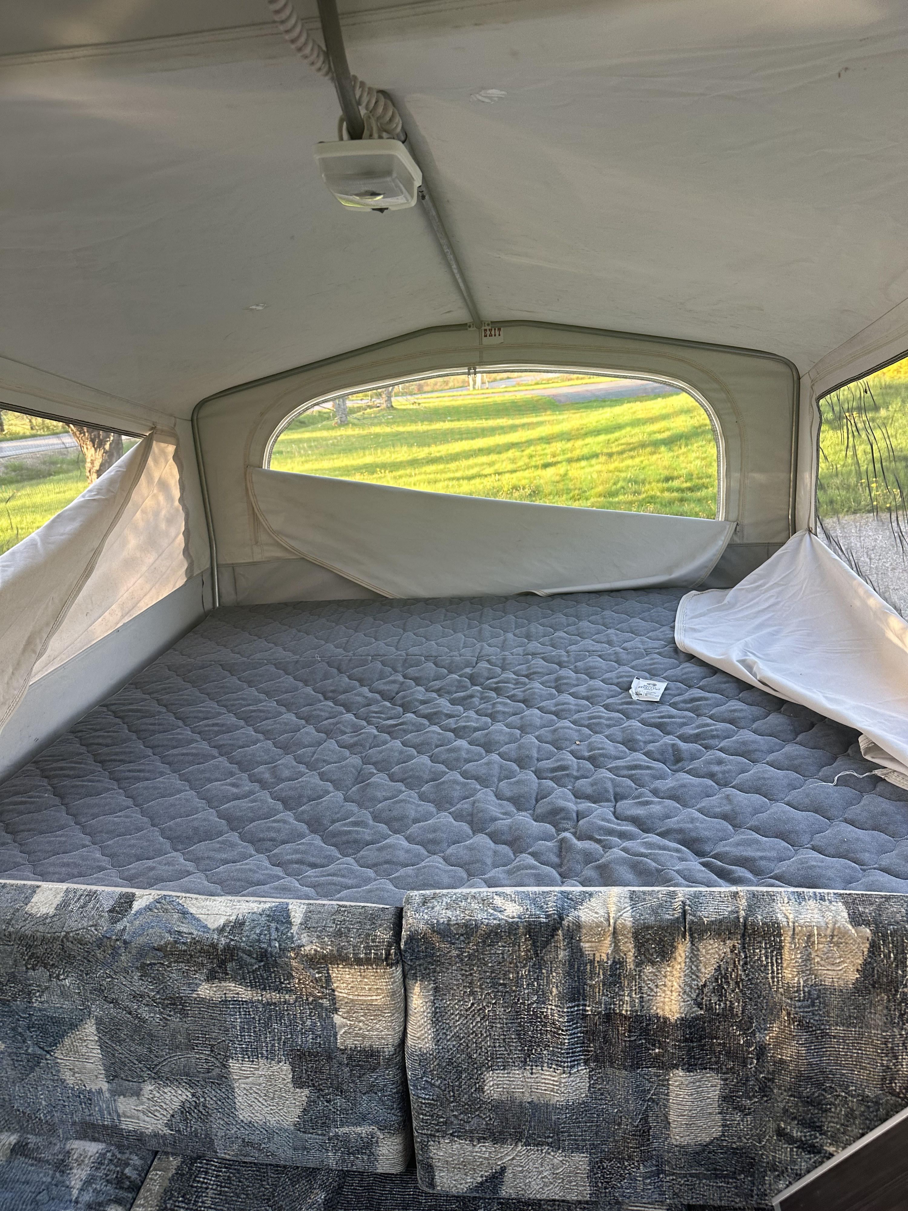 Camping RV rental pop-up interior: comfy gray bed, patterned seating, bright window view of green grass.. Jayco Qwest 2004