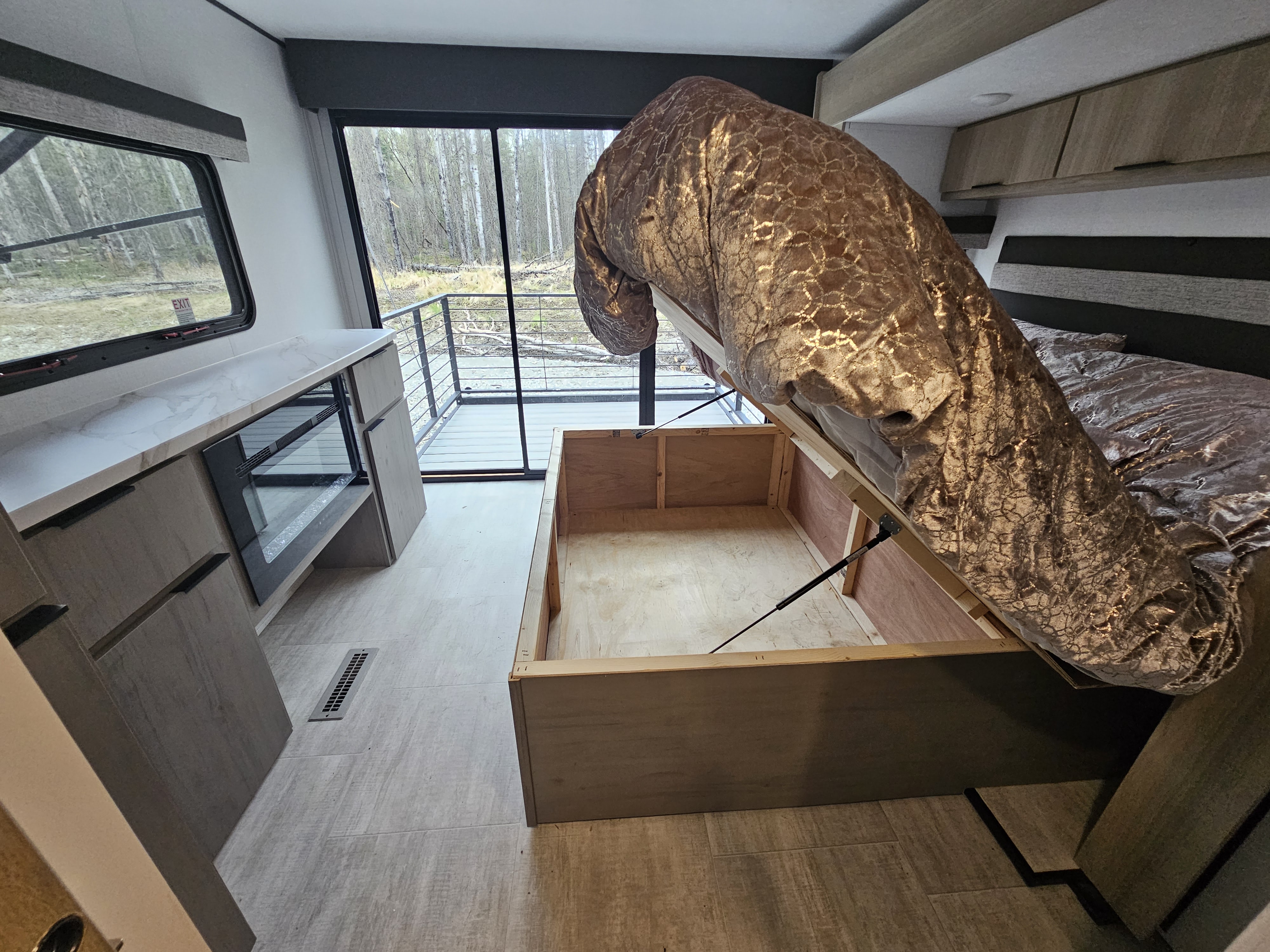 Modern RV rental bedroom featuring a lift-up bed for convenient storage, sleek cabinetry, and large windows with a forest view.. Dutchmen Kodiak Ultimate 2025
