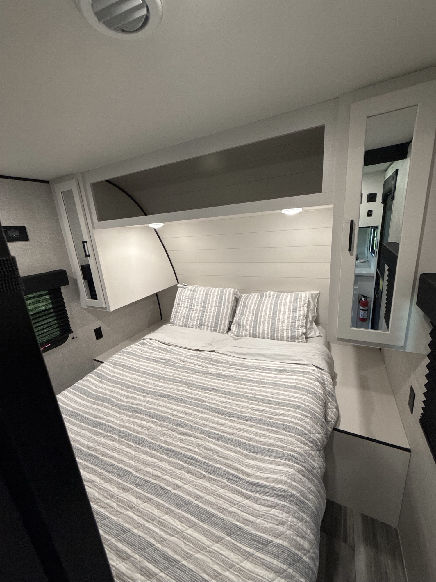 Cozy RV rental bedroom with striped bed, overhead storage, and accent lighting. A mirrored cabinet reflects a fire extinguisher.. Jayco Jay Flight SLX 2024