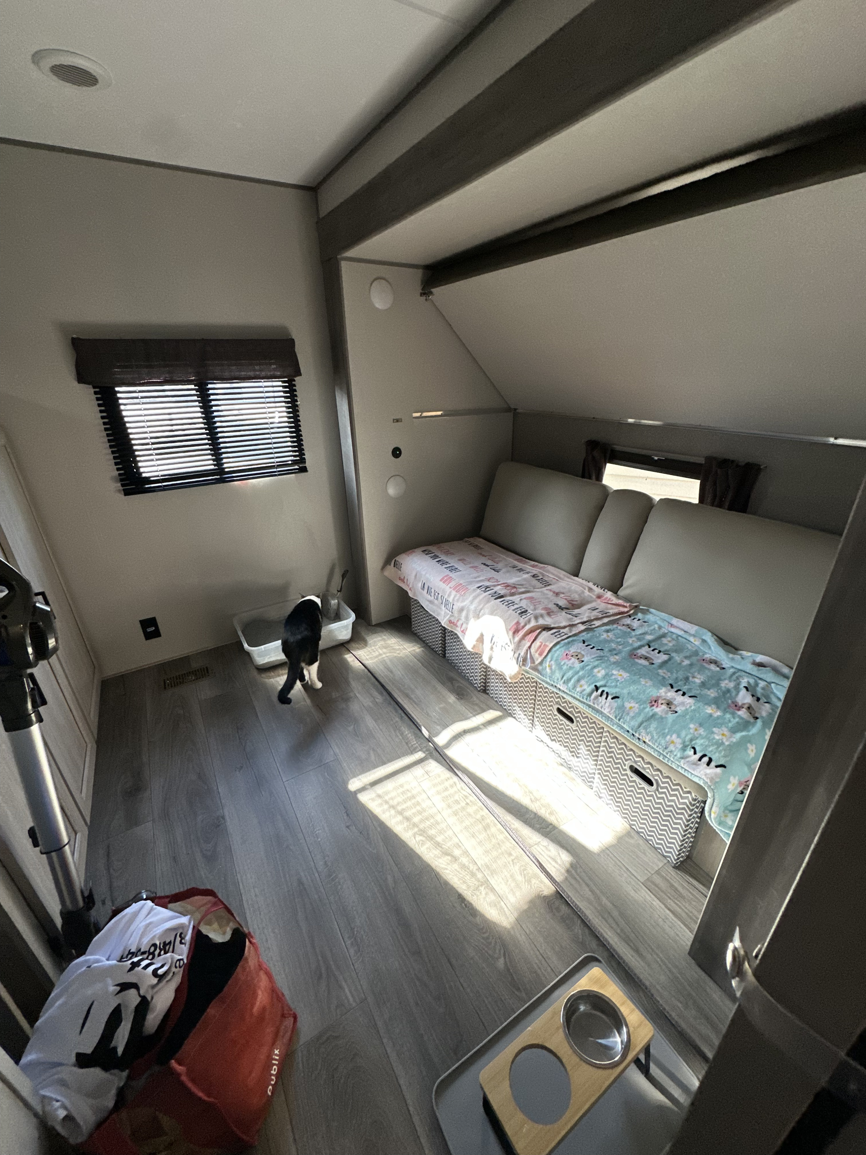 Cozy RV rental interior features a cat by its litter box, a bed with storage, pet bowls, and sunlight on the floor.. Forest River Coachmen Catalina Legacy 2025