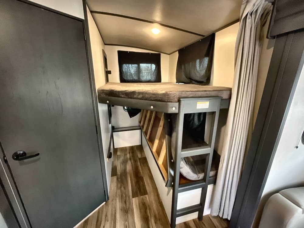 An RV rental's cozy bunk bed sleeping area. Features a ladder, wood-look floor, and windows with blackout blinds for privacy.. Keystone RV Bullet Ultra Lite 2023