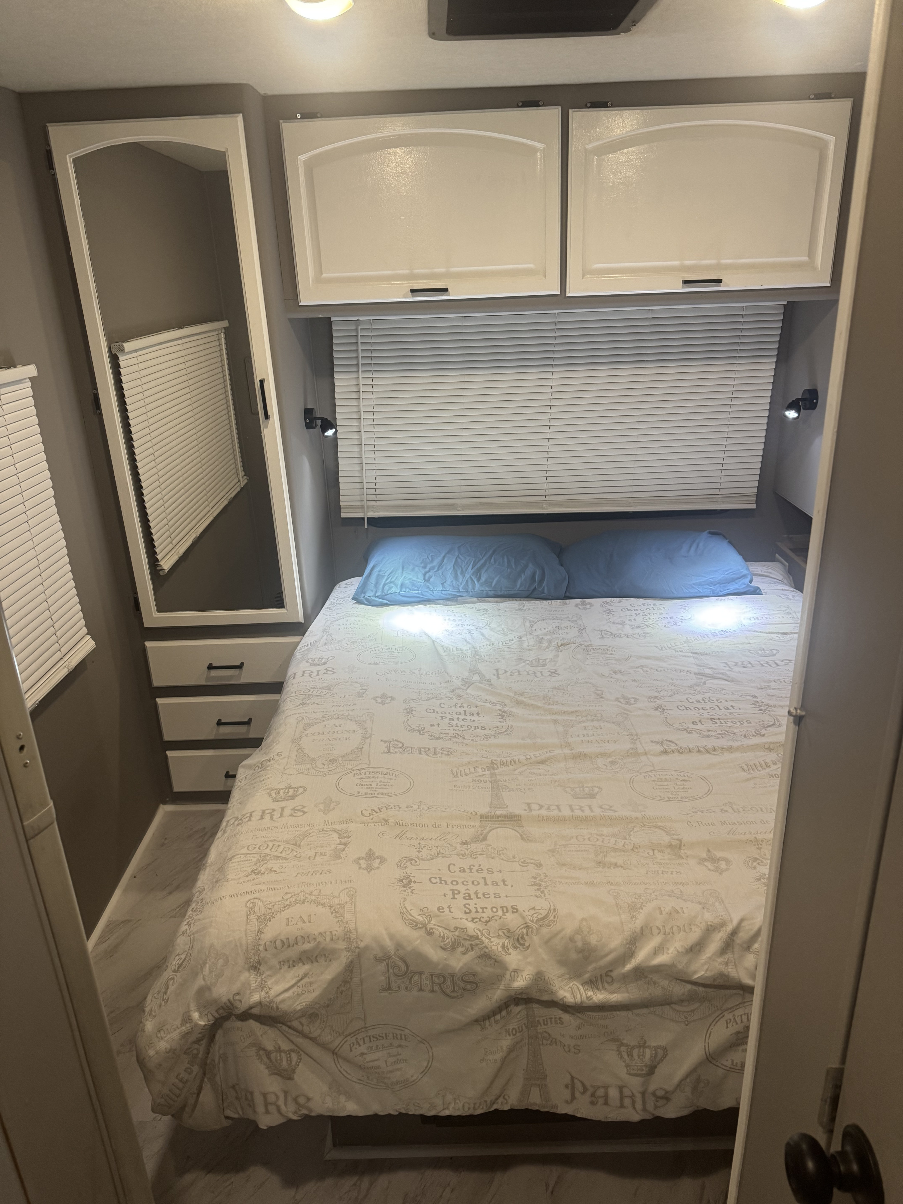 RV rental bedroom with a bed, blue pillows, French-themed duvet, white cabinets, mirror, and window blinds.. Jamboree GT 2010