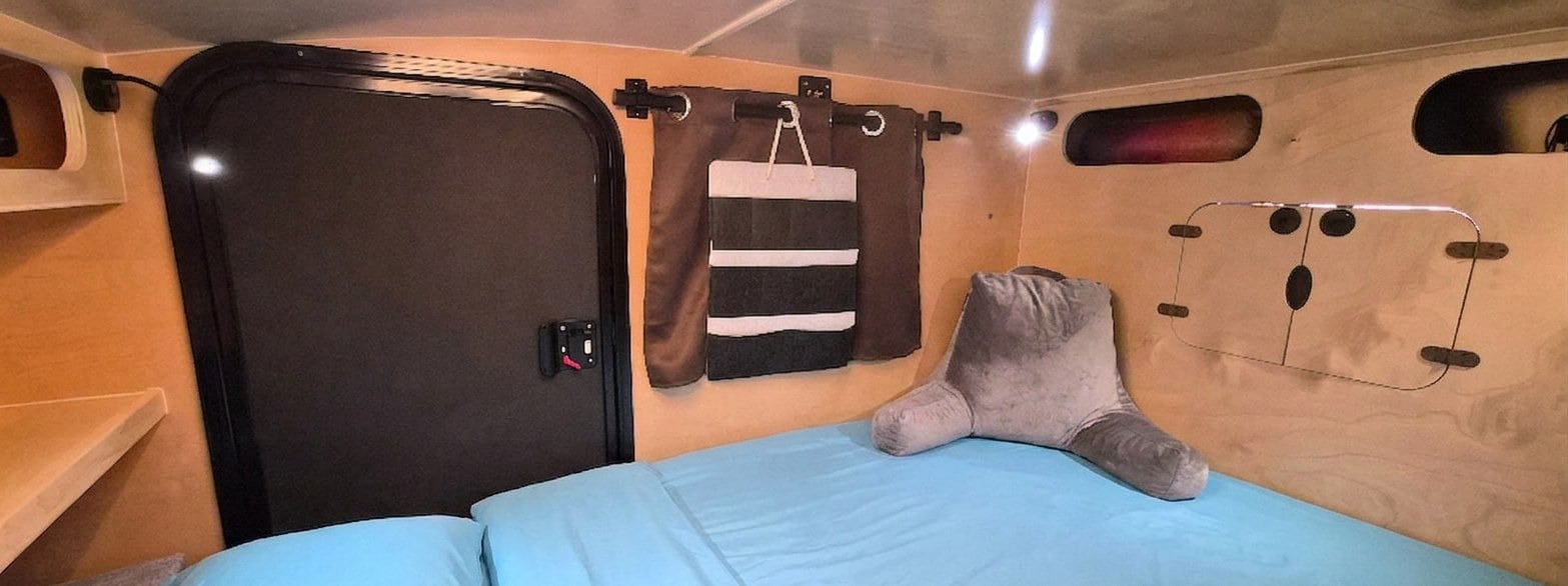 RV rental interior: cozy bed with back pillow, wood paneling, storage cubbies, and a door for your next camper adventure.. Hiker Trailer Backcountry Trailer (Mid Range XL) 2022