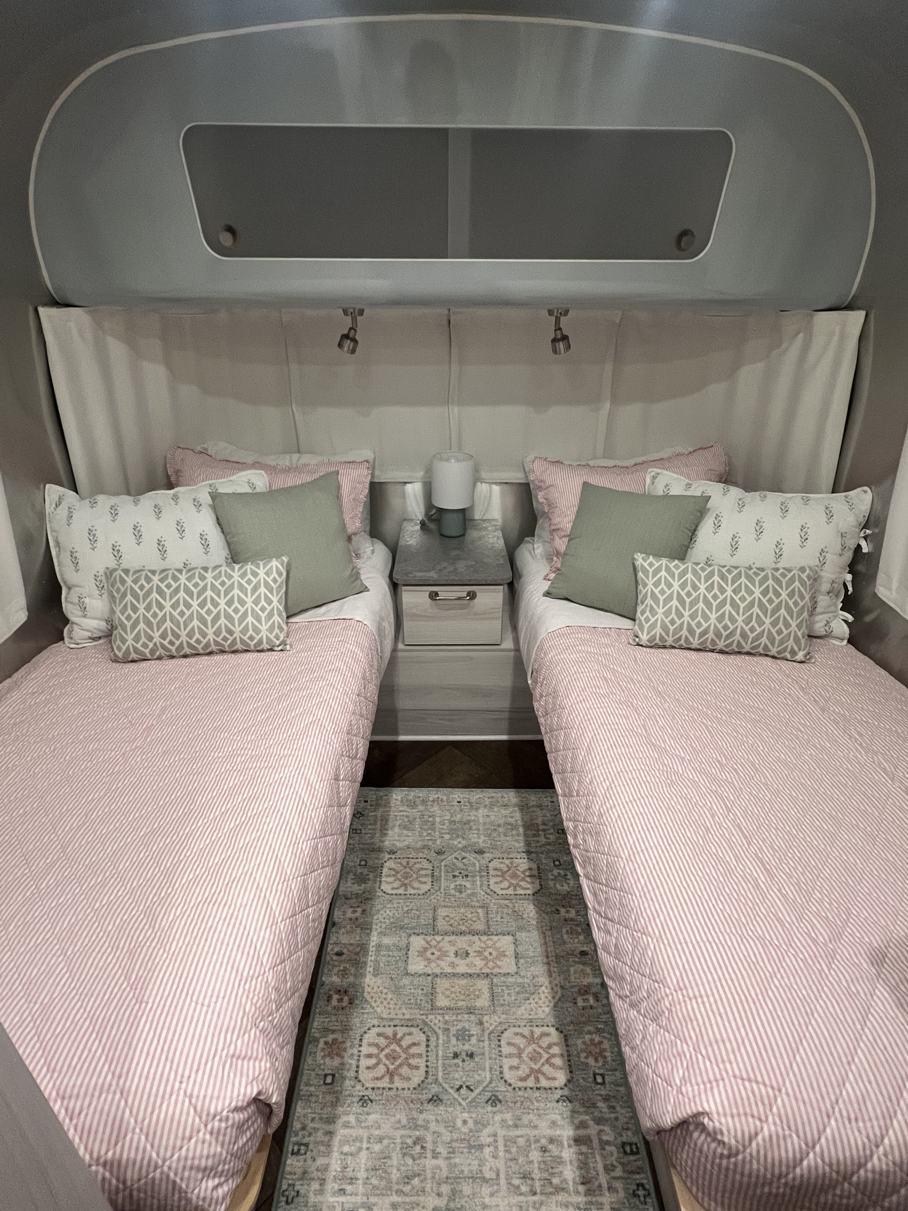 Cozy RV rental bedroom featuring two twin beds with pink striped bedding, decorative pillows, a nightstand, and a patterned rug.. Airstream International 2023
