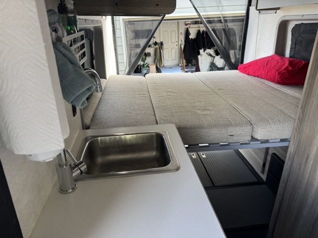 An rv rental camper van interior shows a compact kitchen with sink and paper towels, next to a comfortable bed. Rear open to a garage.. Winnebago Winnebago 2025