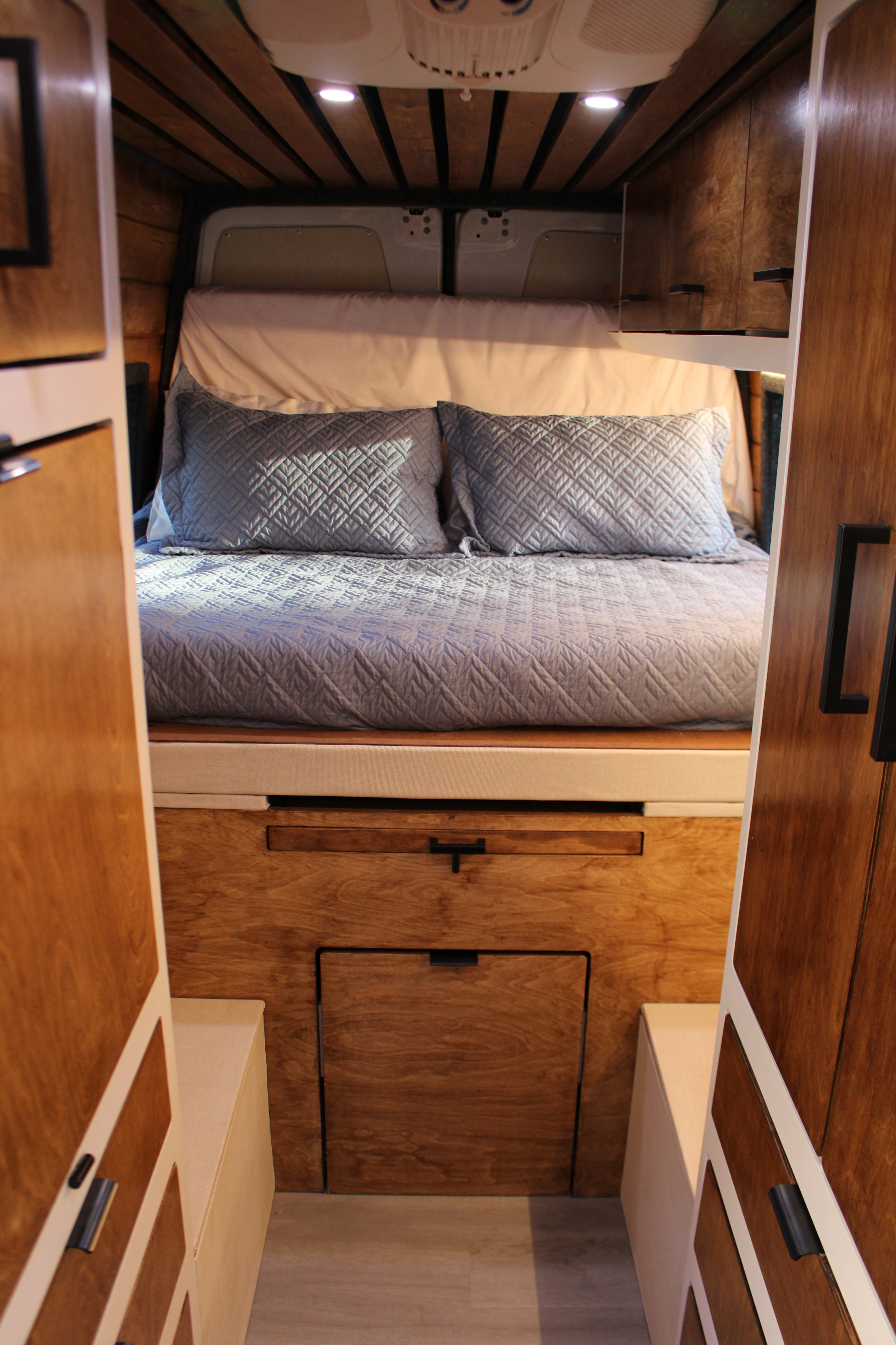 Plush bed in a cozy RV rental, featuring warm wood cabinetry, overhead lighting, and smart storage.. Mercedes Sprinter 2021