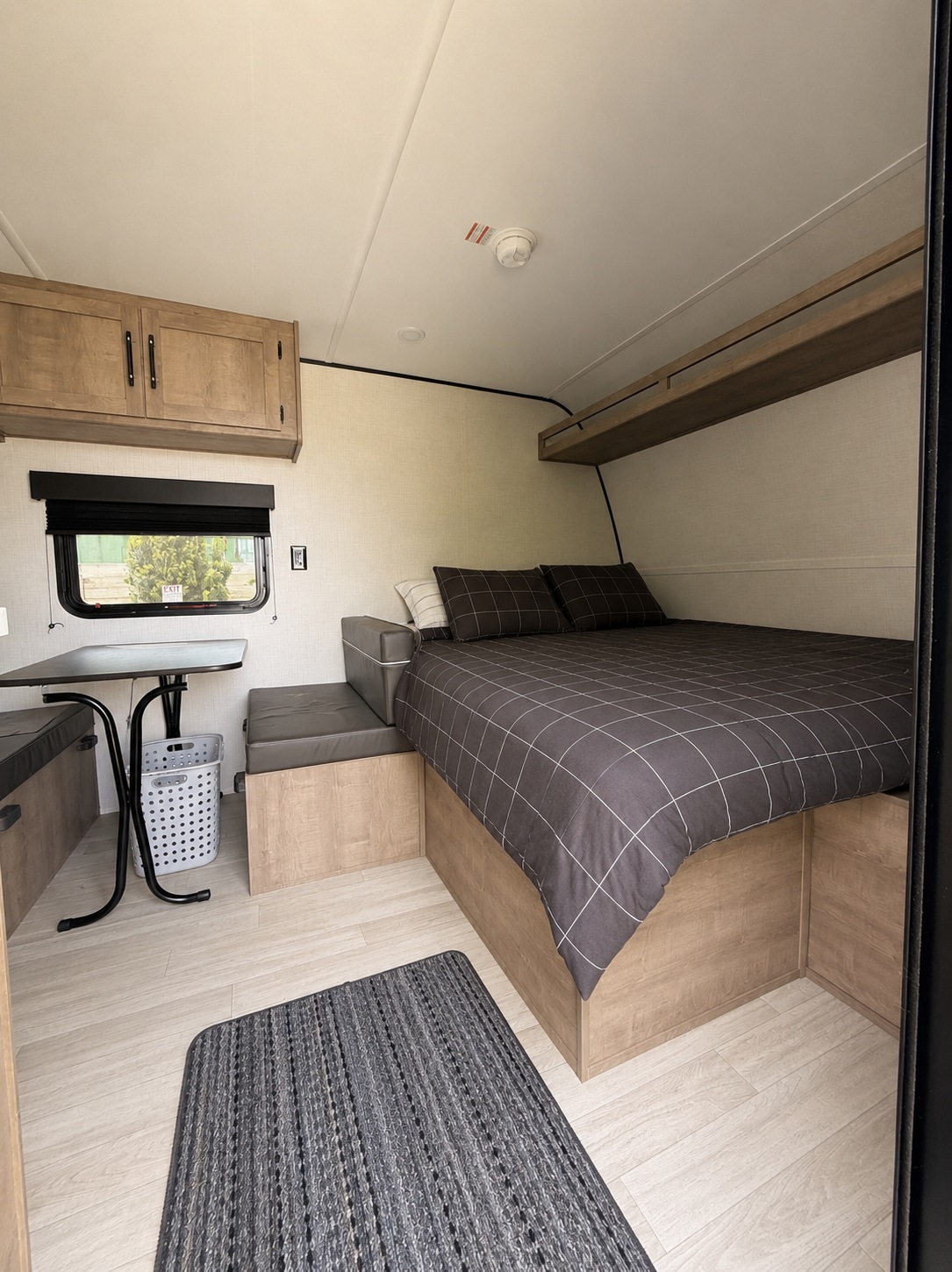 RV rental interior: cozy bedroom with dark checkered bed, overhead shelf, and a compact dining area next to a window.. Keystone RV Springdale Mini 1610BH 2026