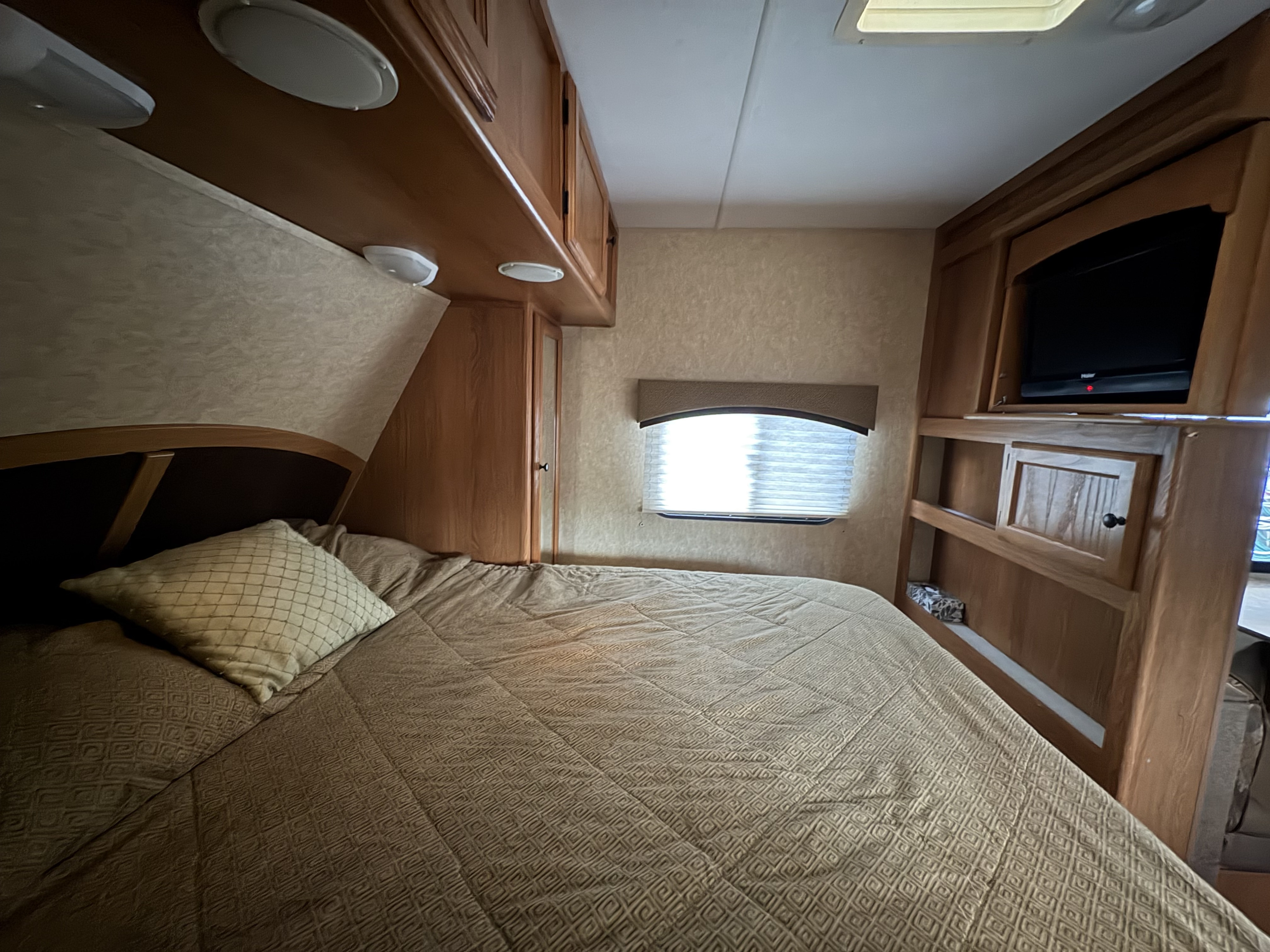 Cozy RV rental bedroom featuring a bed, overhead cabinets, window with blinds, and a mounted TV.. Coleman Lantern Travel Trailer 2011