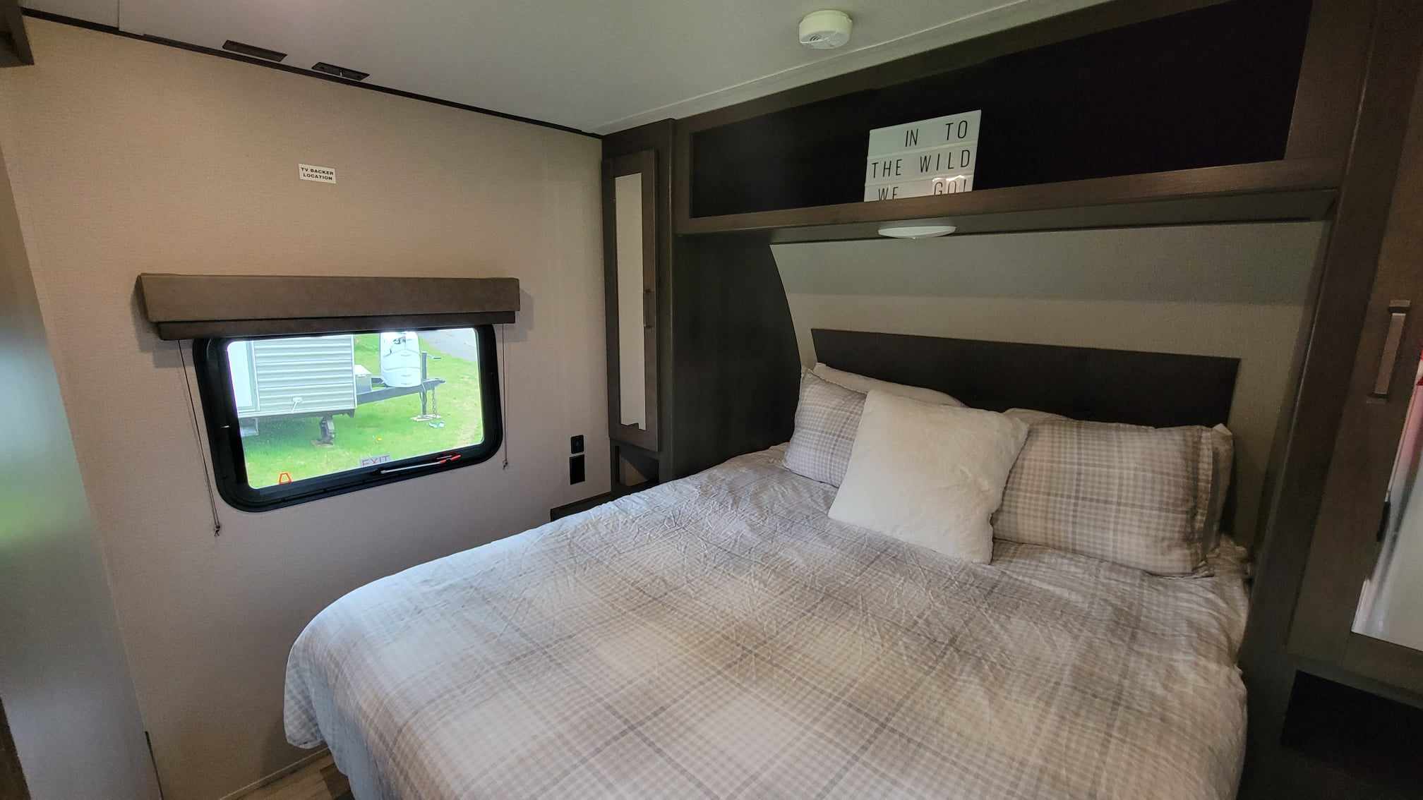 Inside a cozy RV rental bedroom: a comfy bed with plaid linens, window showing outside, and "IN TO THE WILD WE GO!" sign.. Grand Design Transcend Xplor 2022