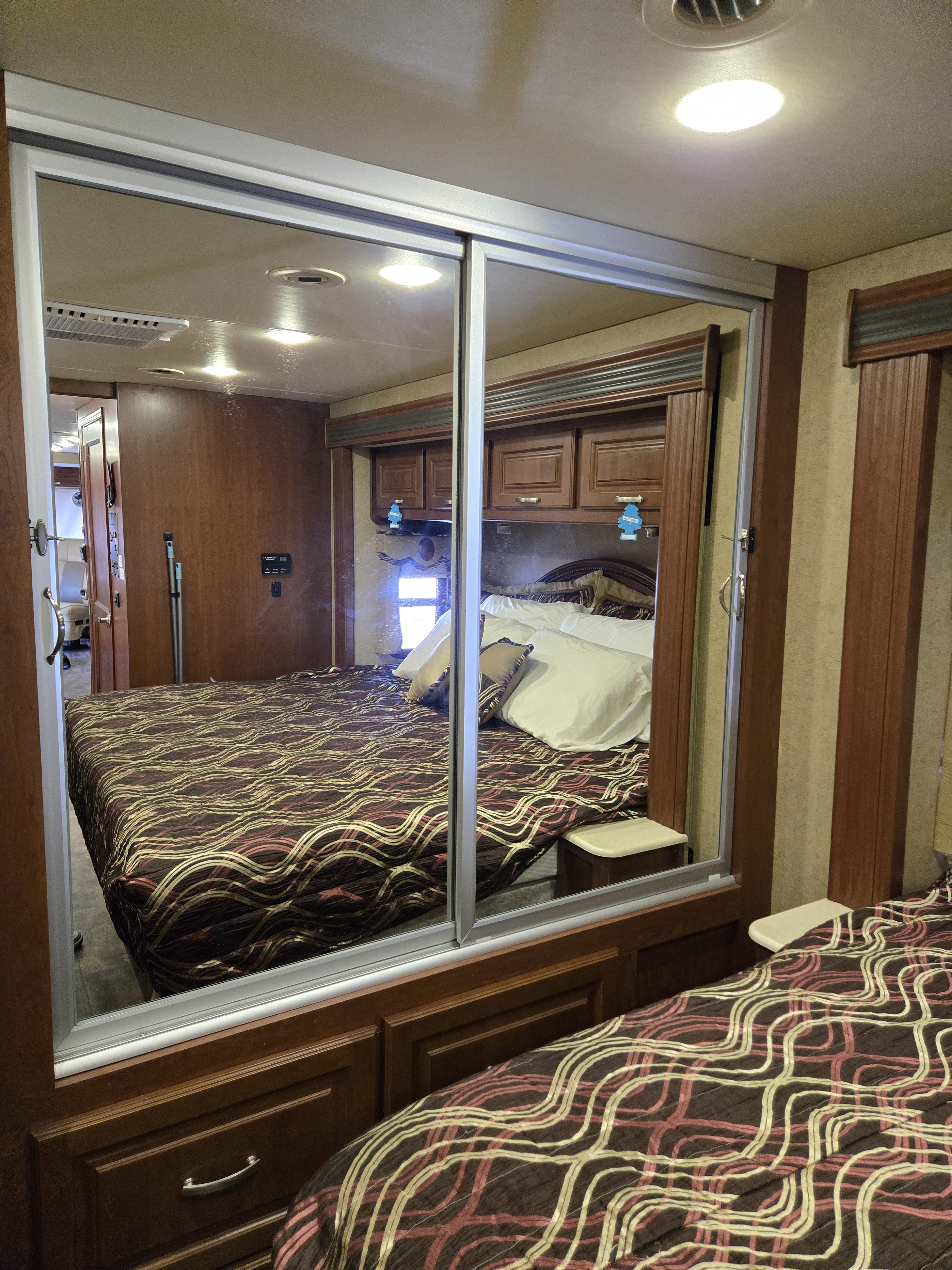 This RV rental bedroom features a large mirrored wardrobe reflecting the bed with a patterned comforter, wood cabinetry, and ceiling lights.. Thor Challenger 2015