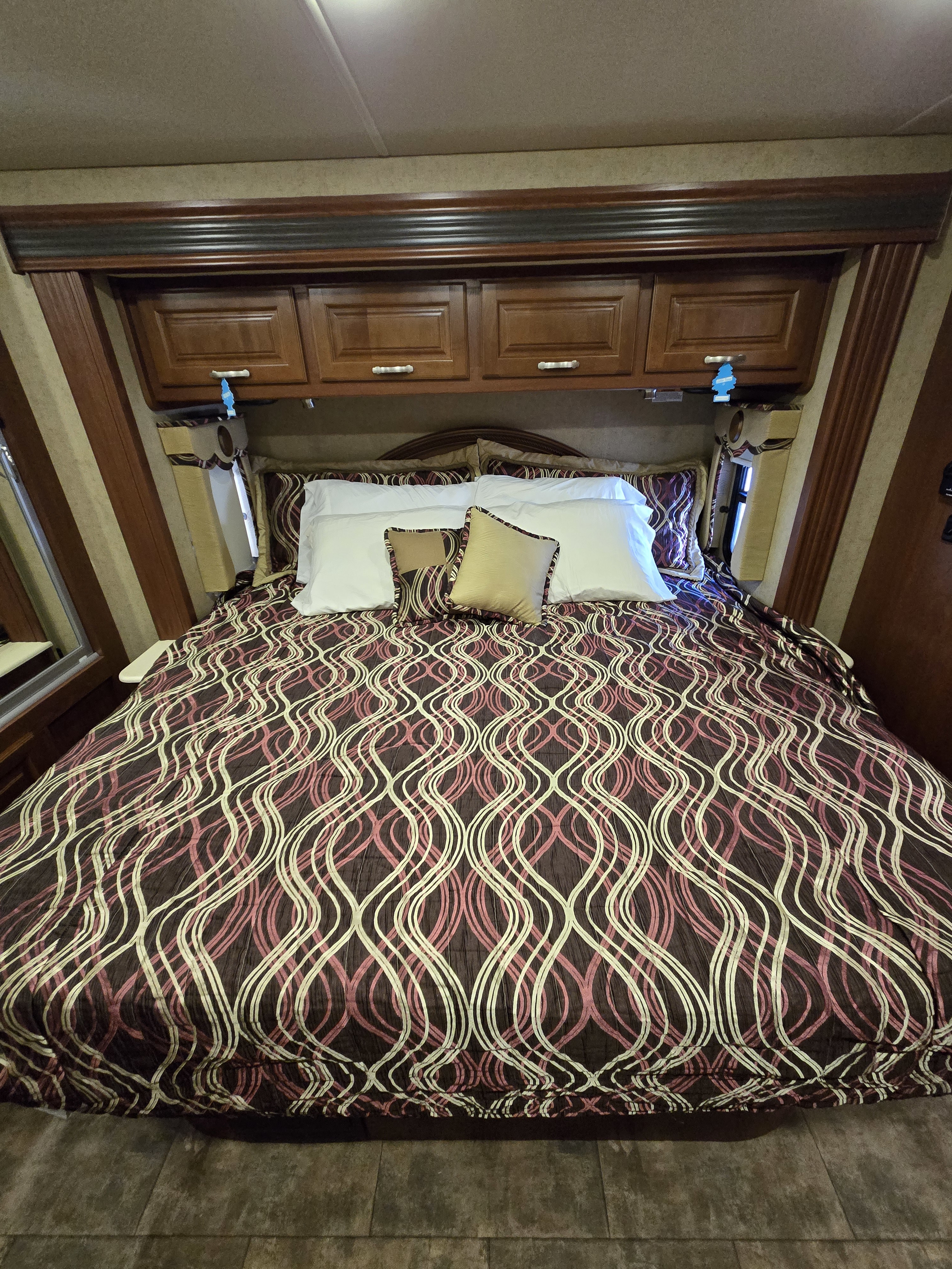 RV rental bedroom featuring a bed with a brown, pink, and cream wavy-patterned comforter, pillows, and wooden overhead storage.. Thor Challenger 2015