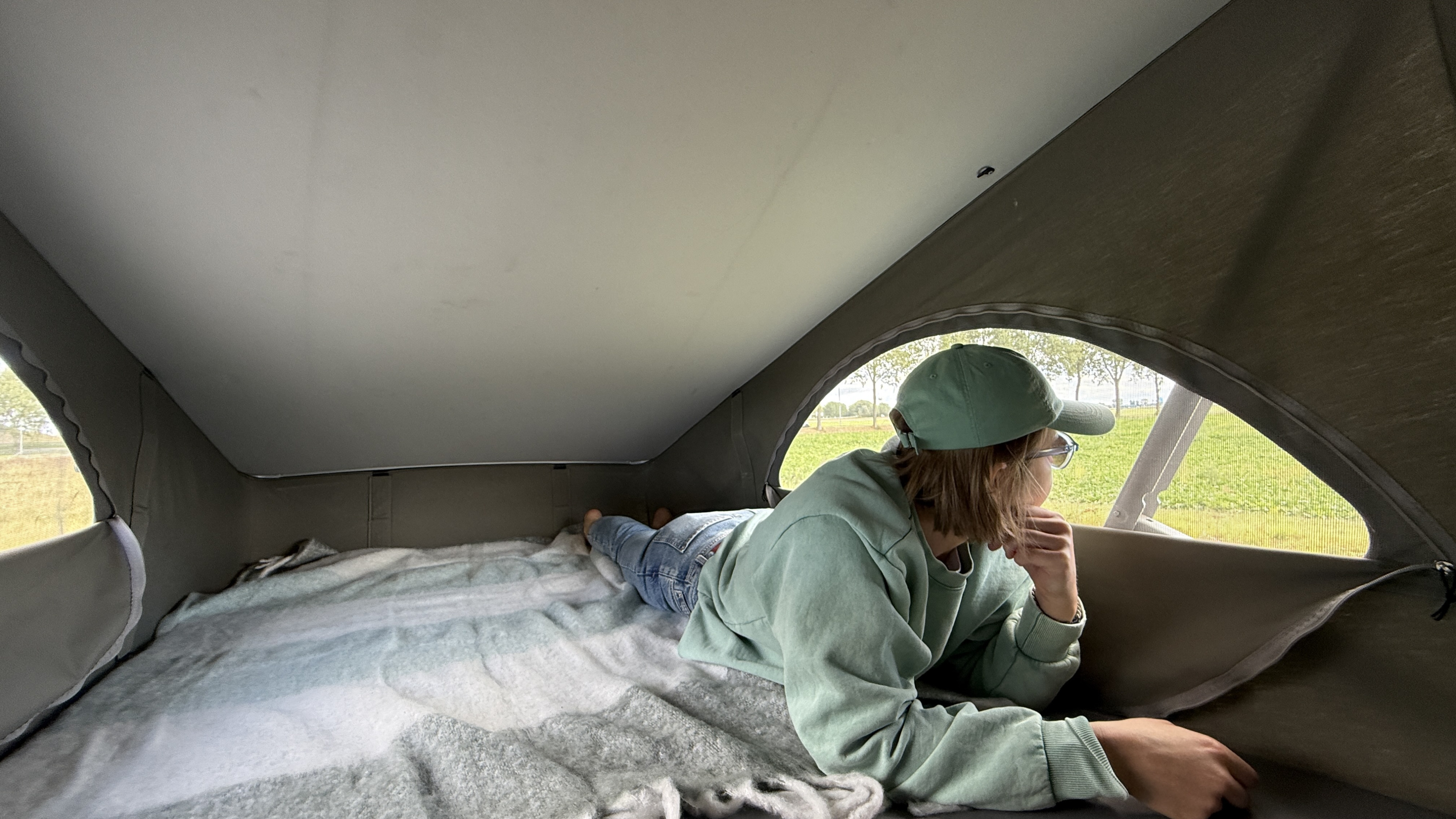Inside an RV rental, a person with a green cap lies on the pop-top bed, looking out at a green field. Relaxing camper travel.. Volkswagen T6 2018