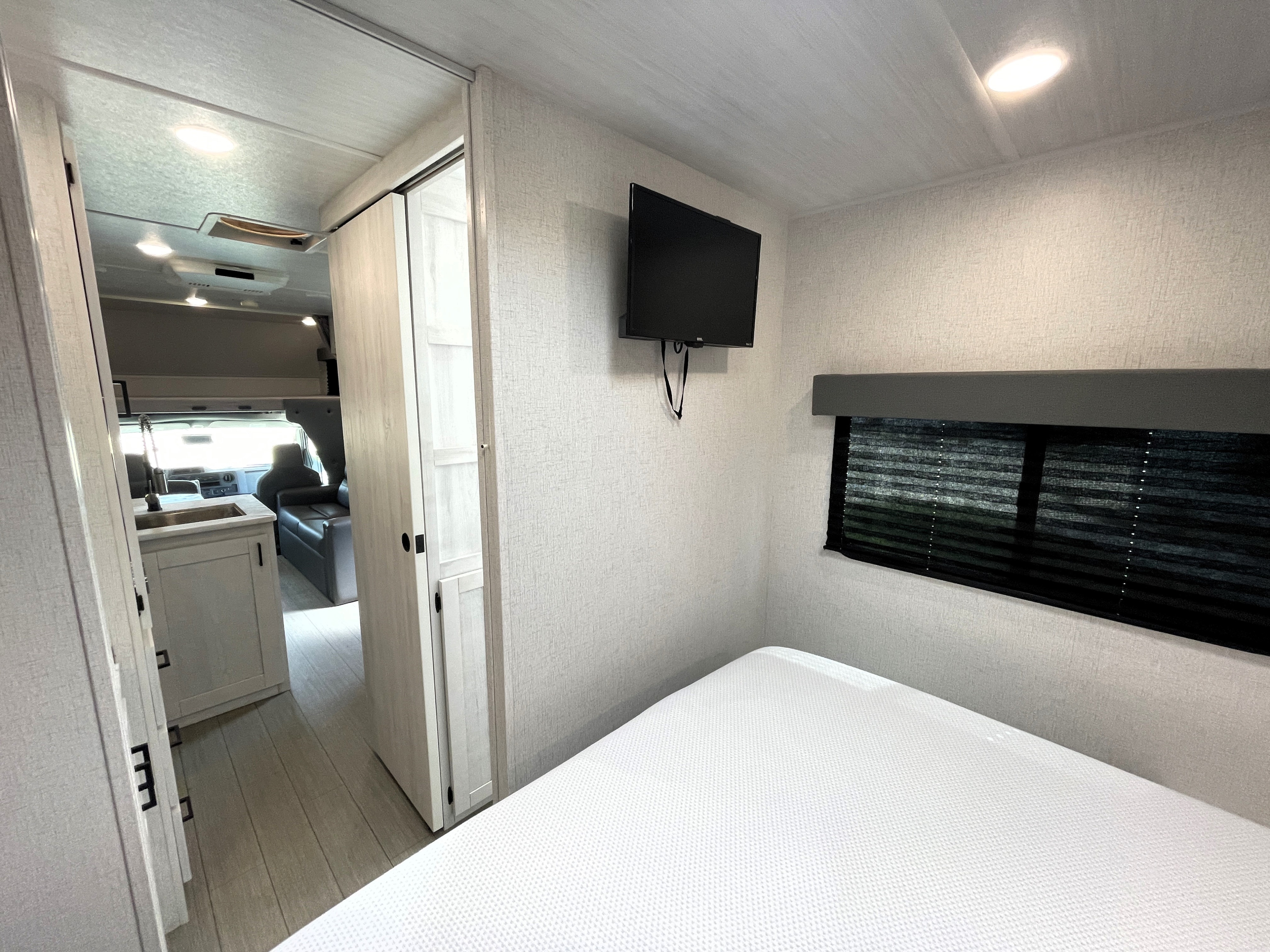 Modern RV rental interior: Bedroom with white bed, wall-mounted TV, and window opens to kitchen & living area with light wood floors.. Entrada Entrada Motorhome 2022