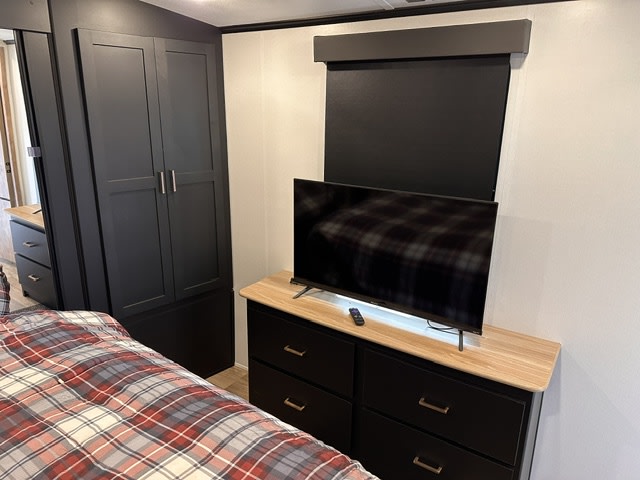 Cozy RV rental bedroom featuring a dark wardrobe, dresser with TV, and a plaid comforter for a comfortable stay.. Keystone RV Outback 2023