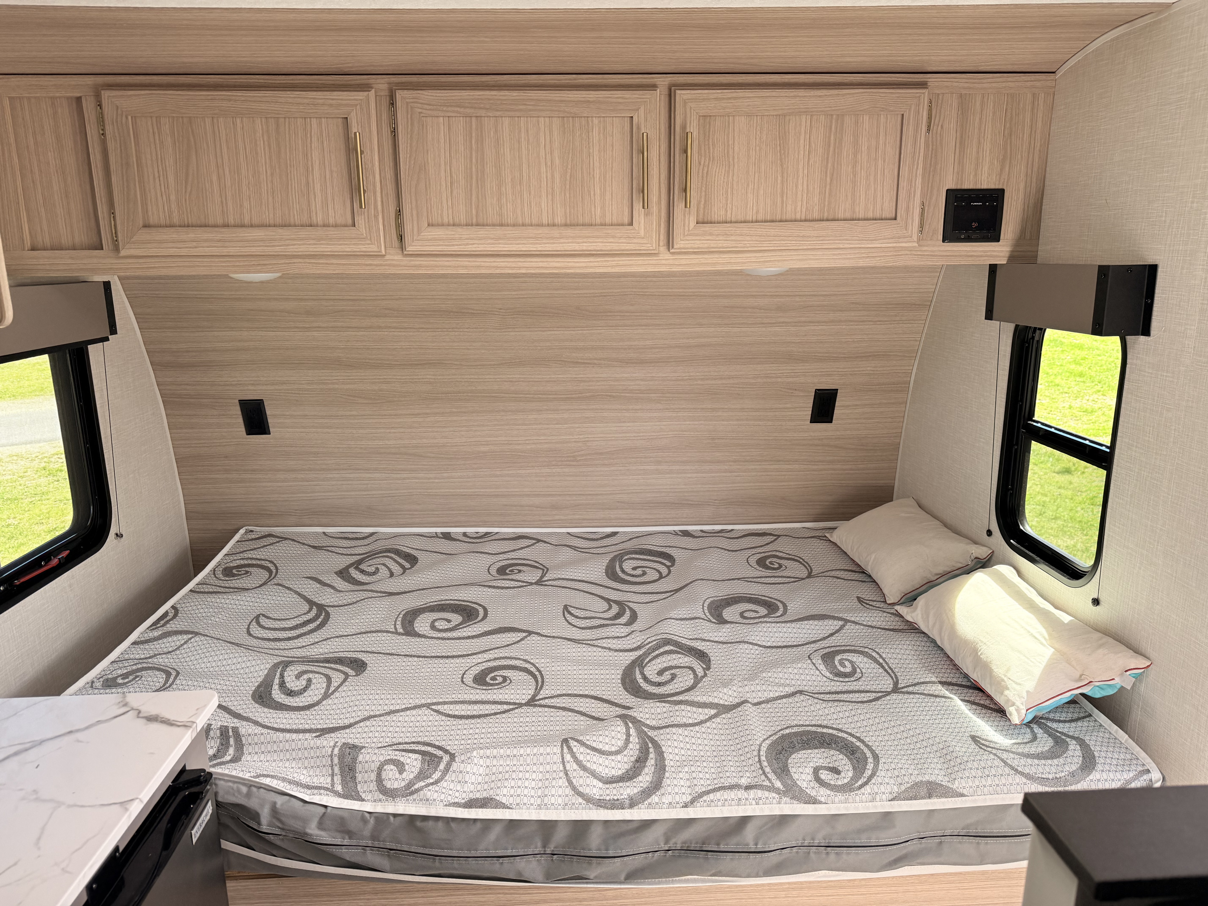 Inside an RV rental: a comfortable bed with a spiral-patterned mattress, light wood overhead cabinets, and bright windows.. KZ Sportsmen Classic 2025