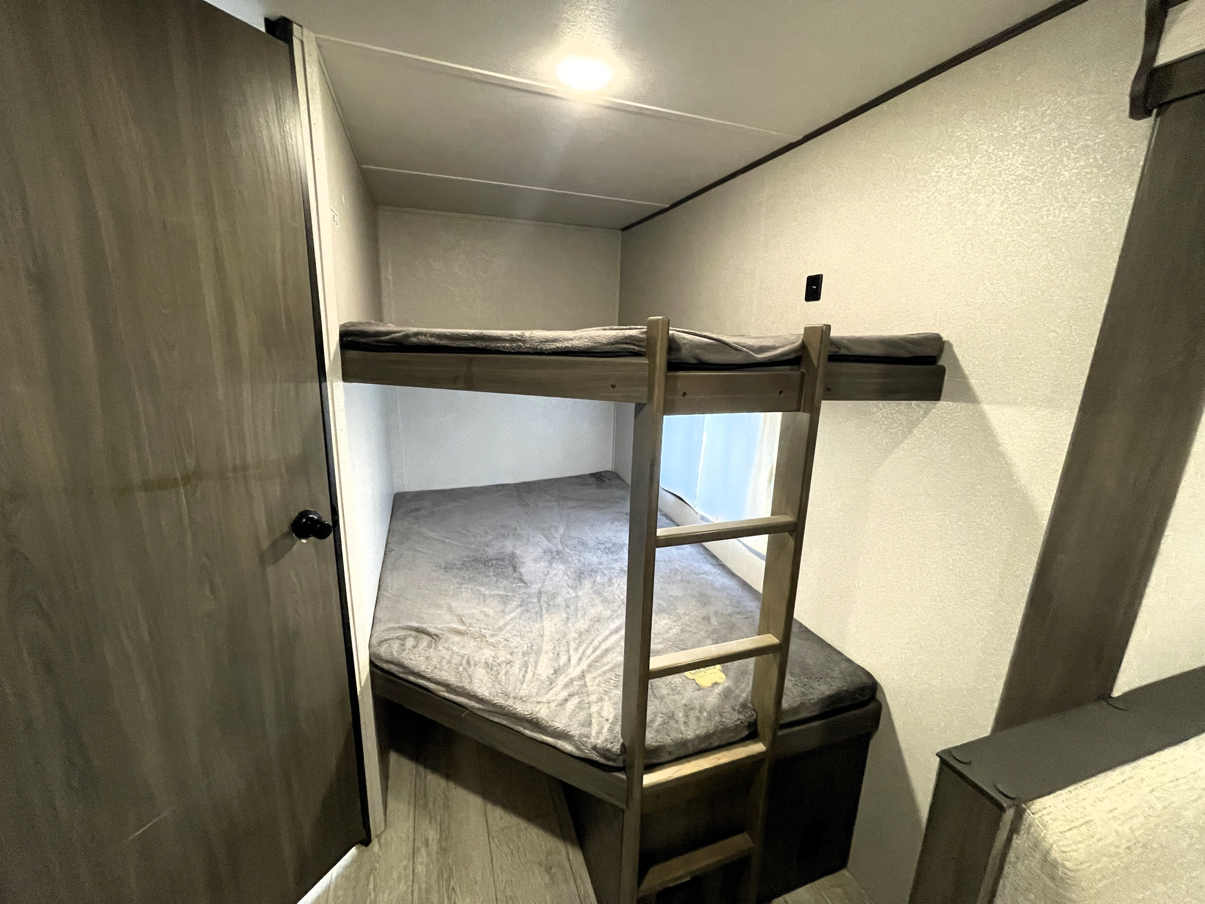 Cozy bunk beds with grey bedding & a wooden ladder in an RV rental. An overhead light illuminates this compact sleeping area.. Shasta 26DB 2021